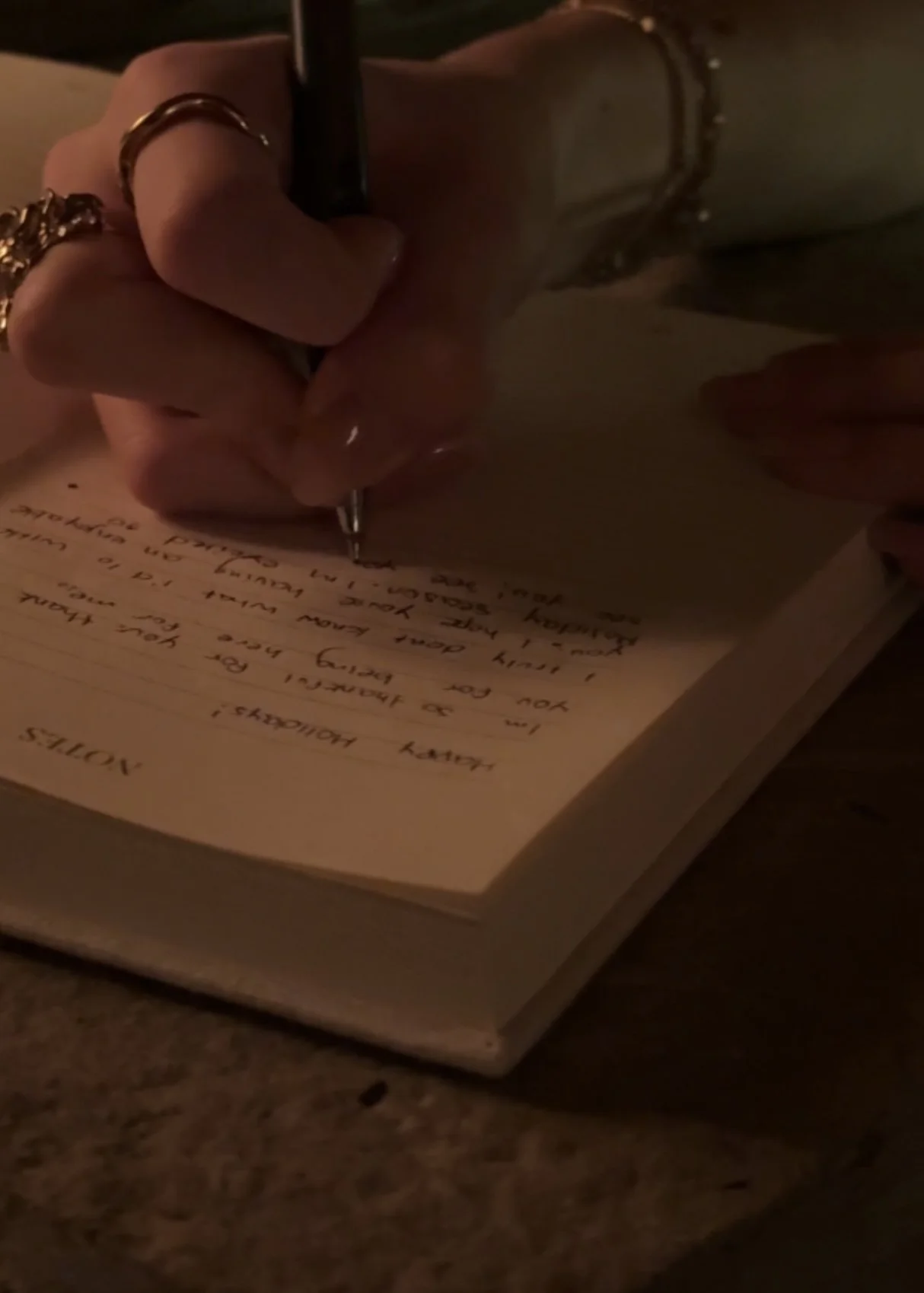 A person with rings and a bracelet writing in a notebook with a pen under dim lighting.