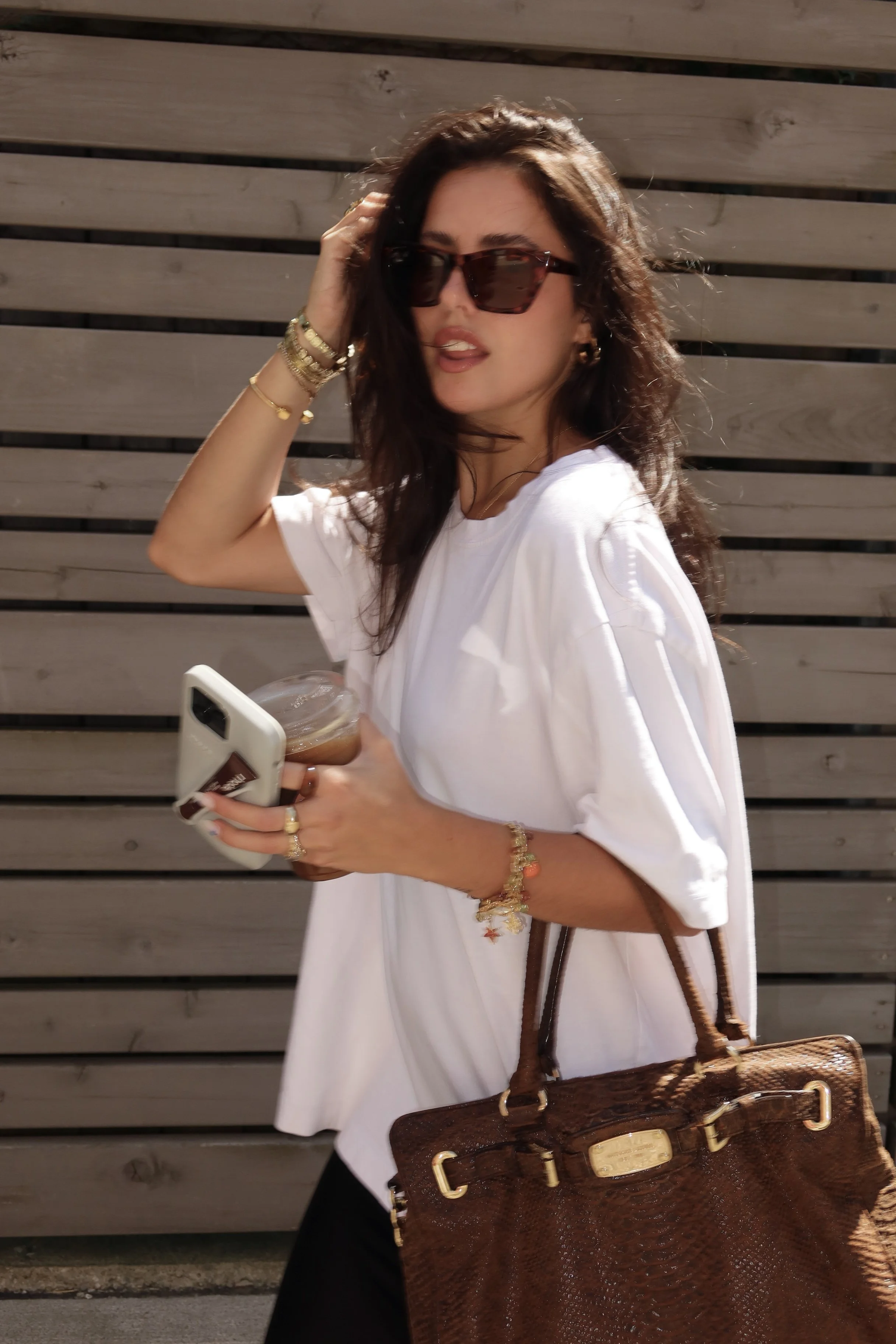 A woman with dark hair wearing sunglasses, white t-shirt, gold jewelry, carrying a brown handbag, holding a phone and a drink, standing in front of a wooden fence.