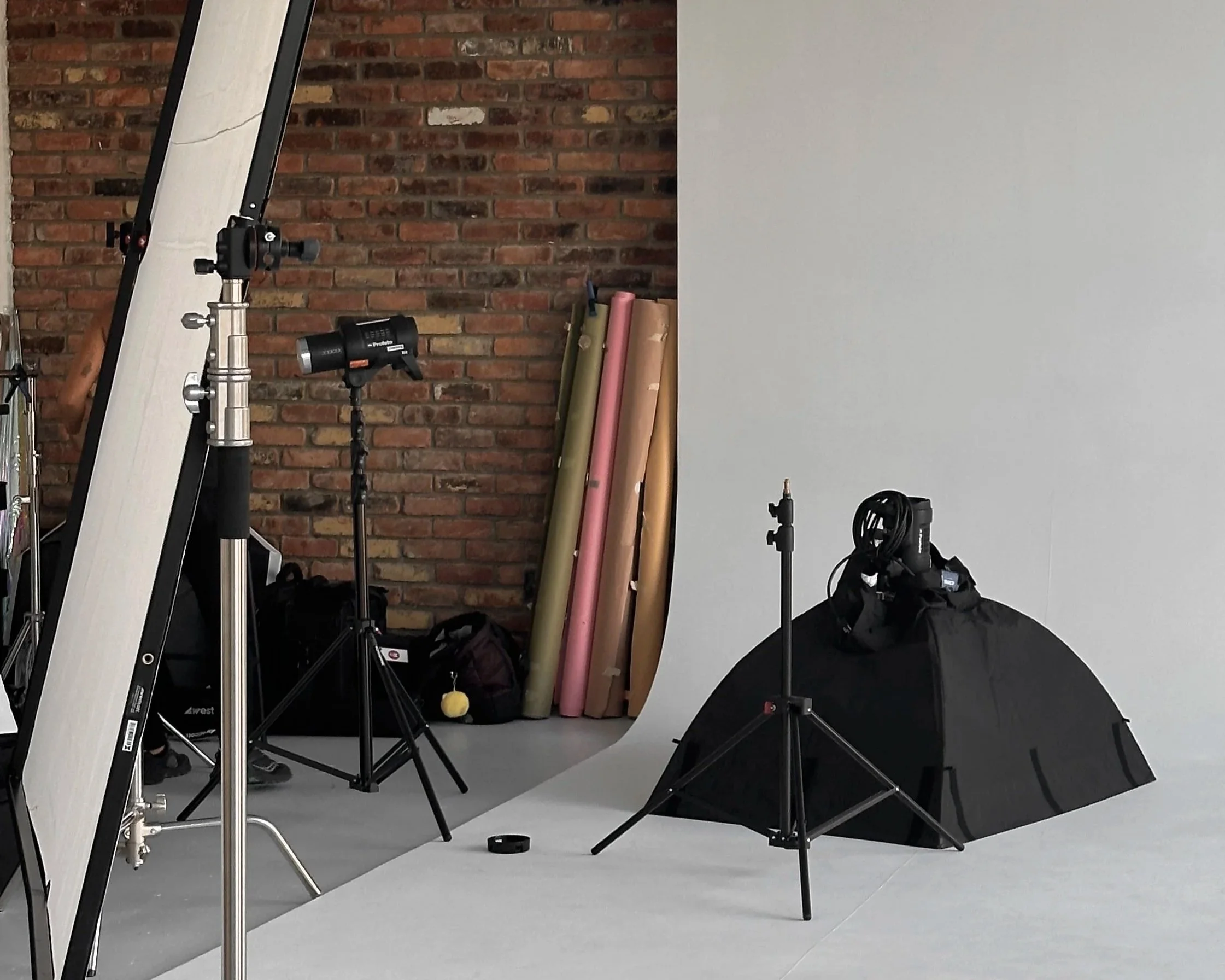 Photography studio setup with tripod, lights, backdrop, and colored paper rolls against a brick wall.