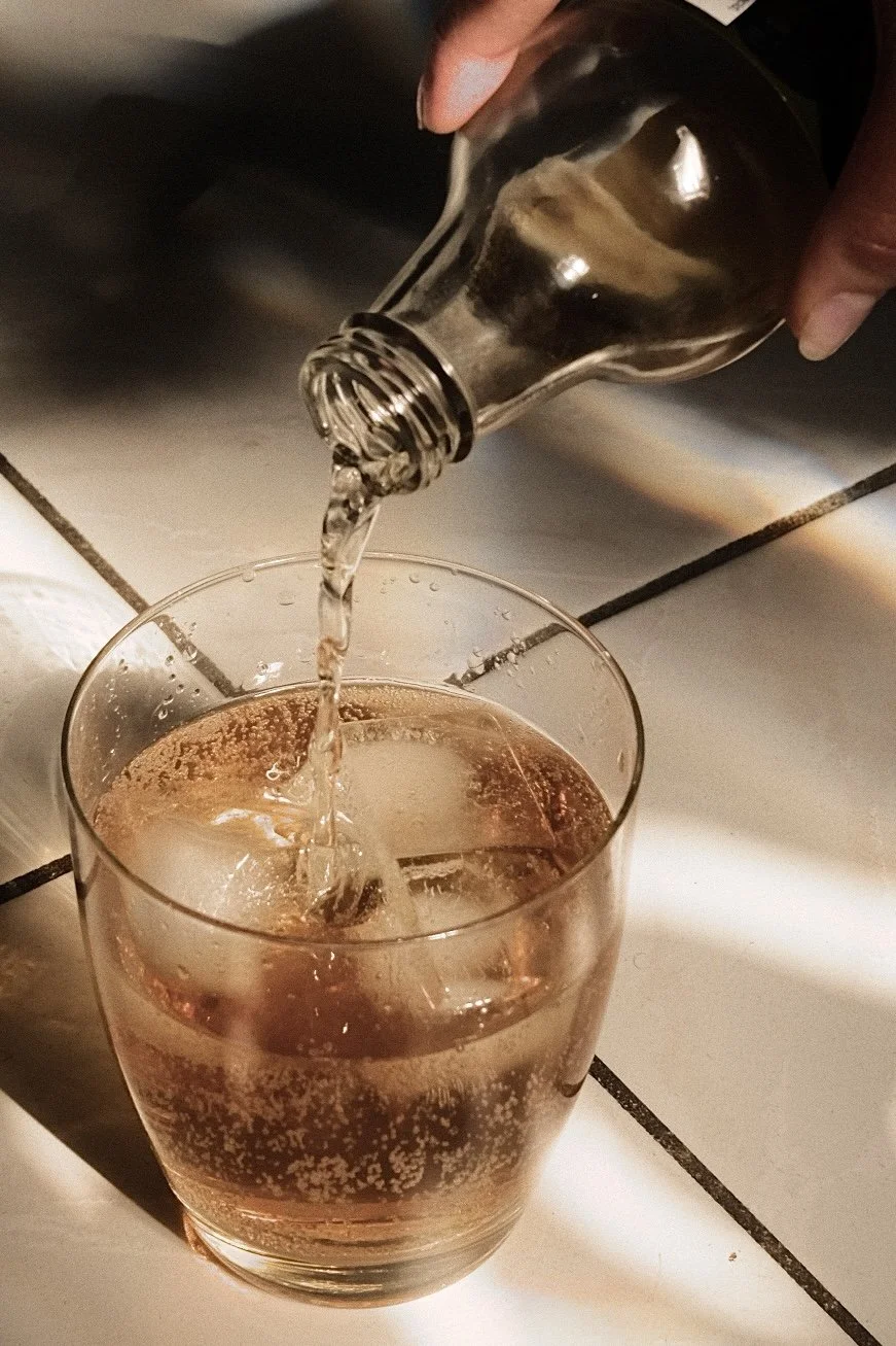 Person pouring clear soda into a glass over a tiled countertop.