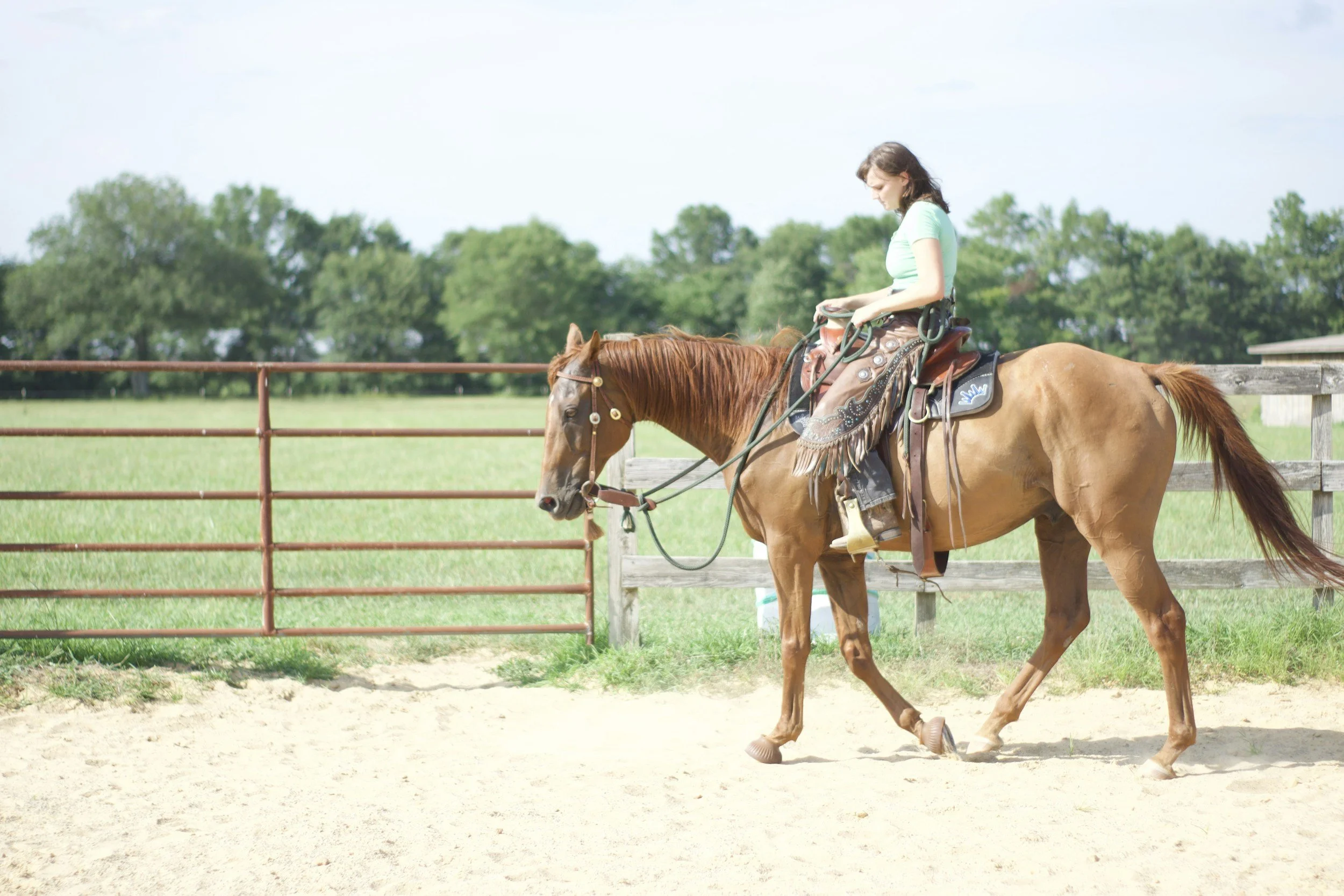 A woman riding a brown horse in an outdoor arena on a sunny day. She is wearing a light green top and jeans, with the horse equipped with western riding gear.