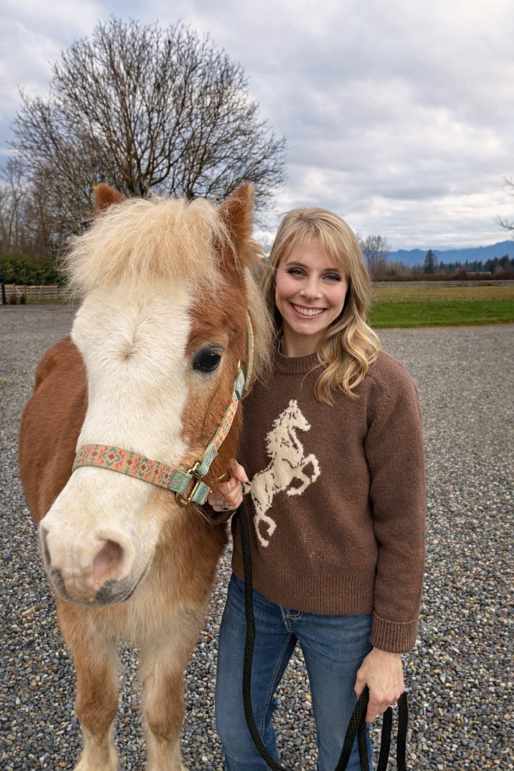 A woman in Enumclaw with blonde hair smiling and standing next to a brown and white horse outdoors on a cloudy day.