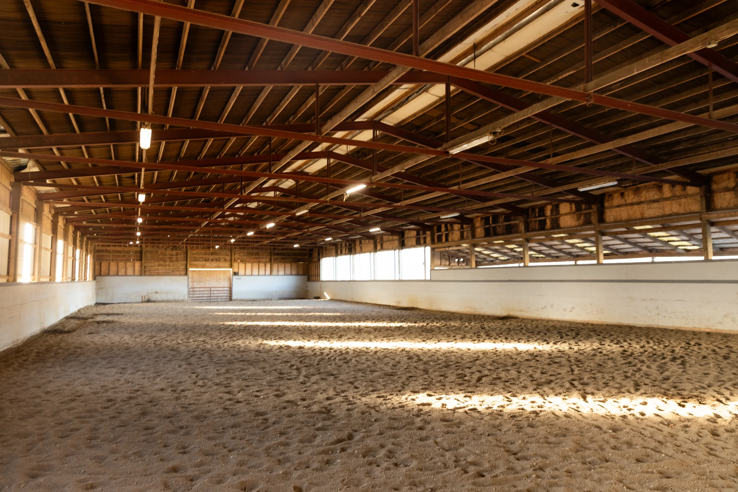 Interior of an indoor horse riding arena in Enumclaw with a sandy floor, wooden walls, and a high ceiling with exposed beams and lighting.