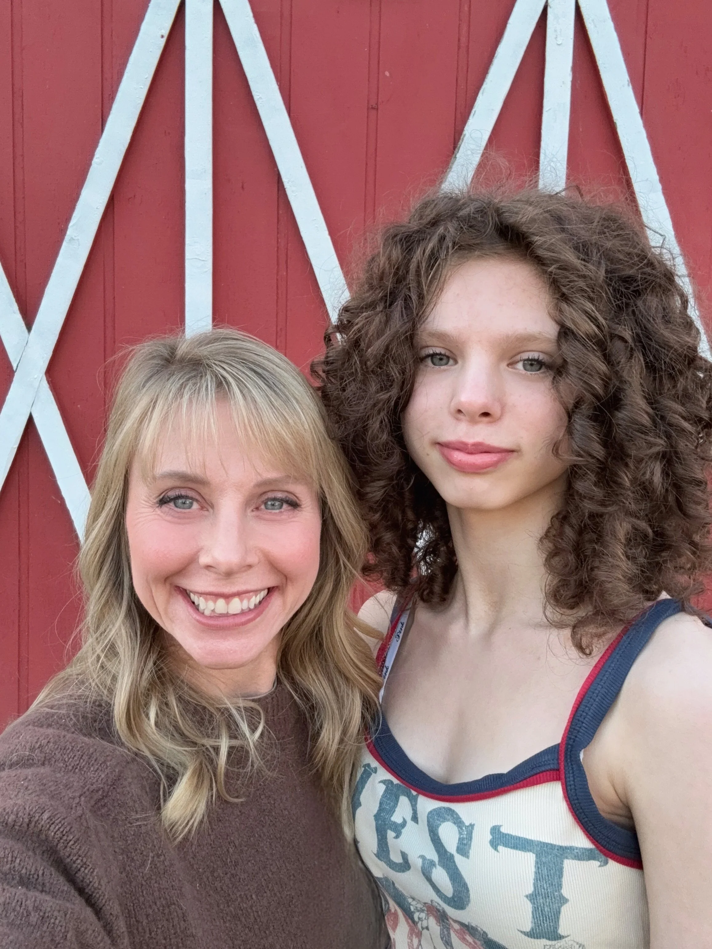 Two women in Enumclaw taking a selfie in front of a red barn with white trim. The woman on the left has blonde hair, is smiling, and wearing a brown sweater. The woman on the right has curly brown hair, is wearing a tank top with text on it.