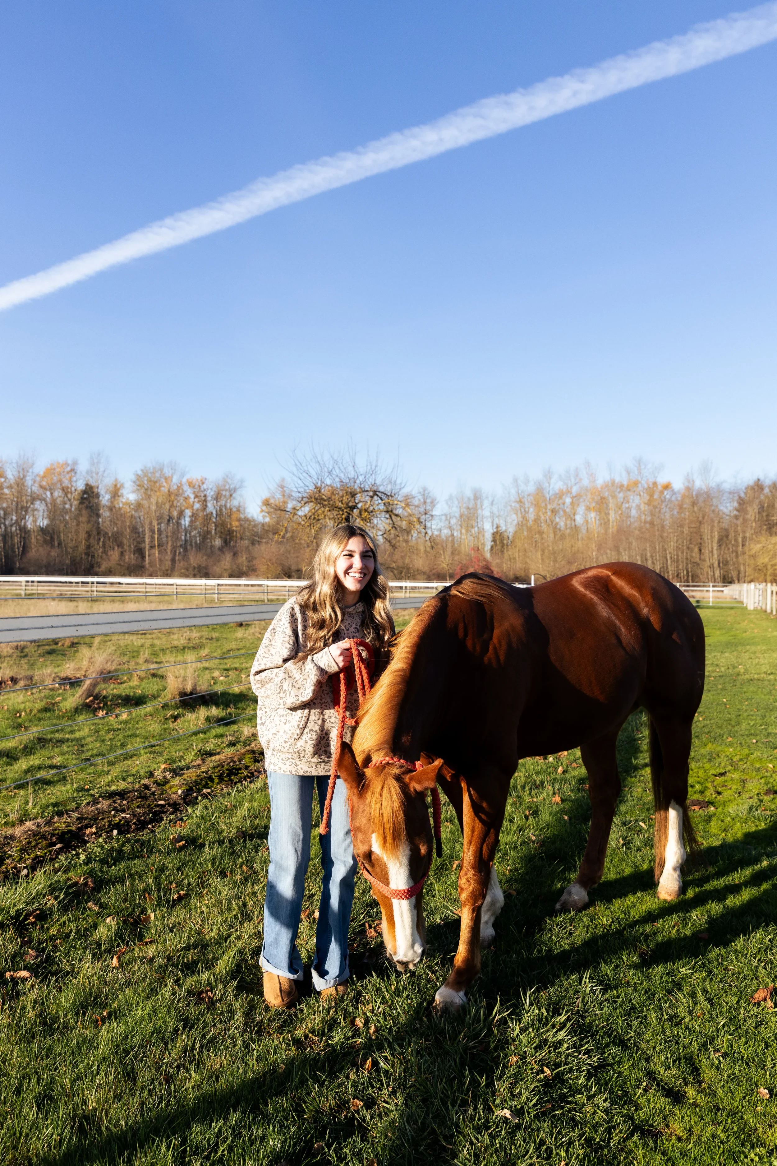 A young woman with long, wavy blonde hair standing on green grass next to a chestnut horse on a farm or pasture. She is holding the horse's halter while the horse grazes in Enumclaw