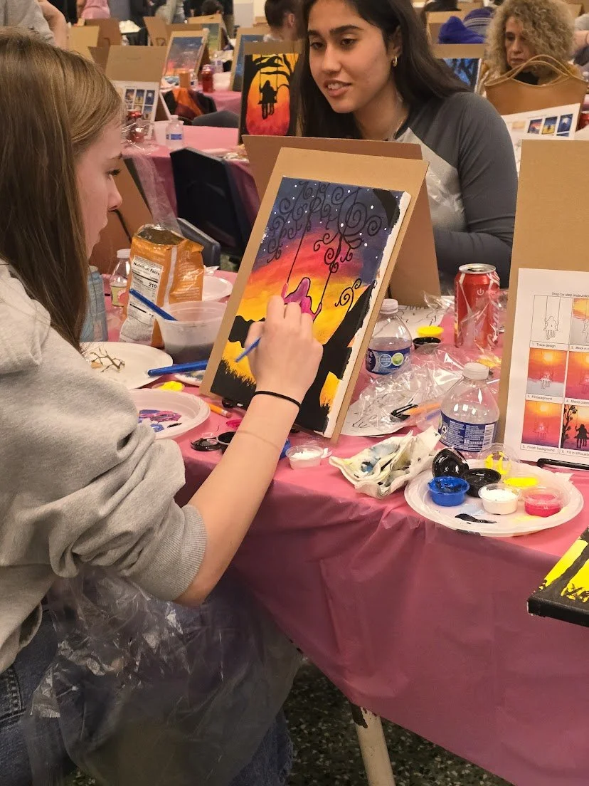 A young woman in Enumclaw paints a sunset scene with a black silhouette of trees and a person on a swing, on a canvas at a painting event. She is seated at a table with painting supplies, including paint palettes, and snacks.