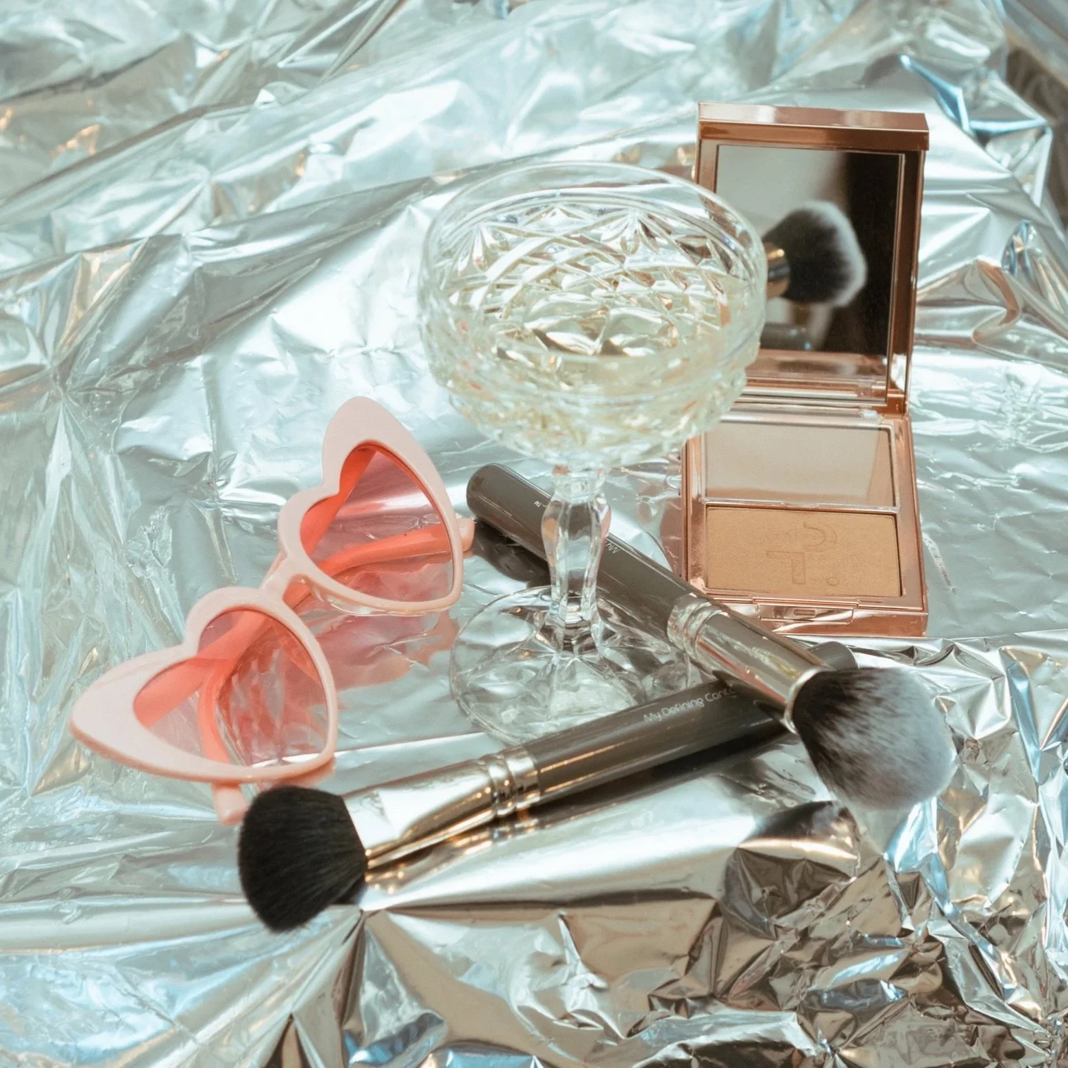 Beauty products including a makeup palette, brushes, a glass dish, heart-shaped sunglasses, and a makeup sponge on a crinkled silver foil background.