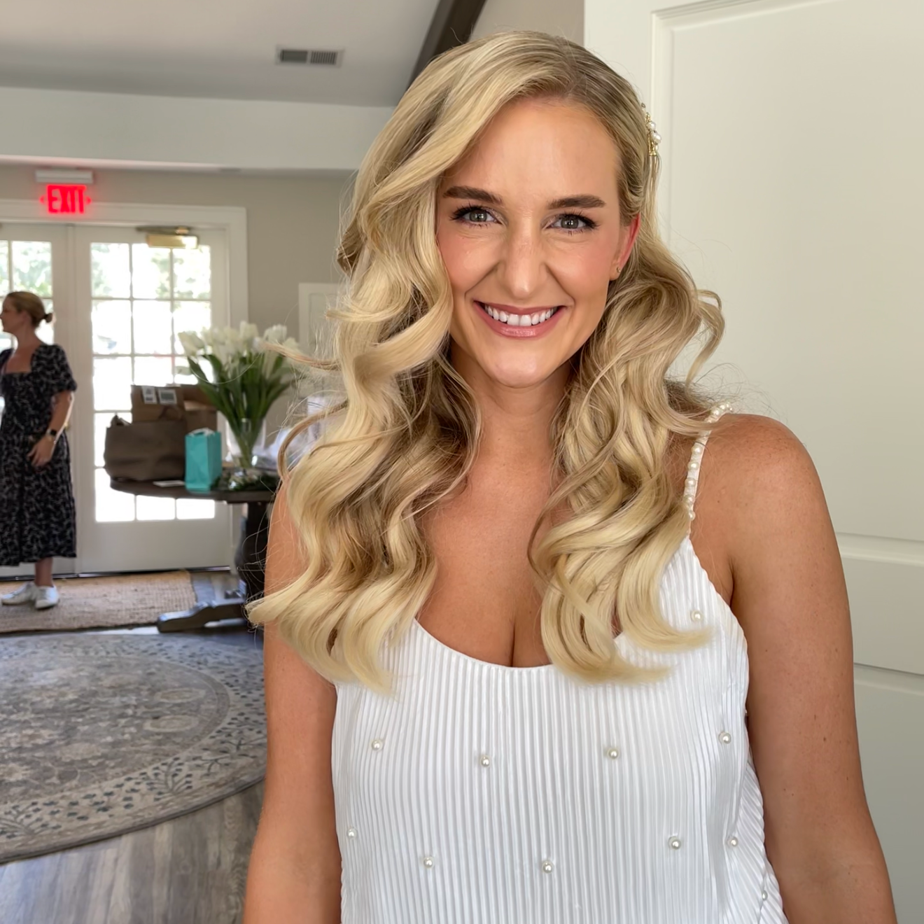 A smiling blonde woman with long, curly hair wearing a white dress with pearl embellishments, standing indoors in a well-lit room.