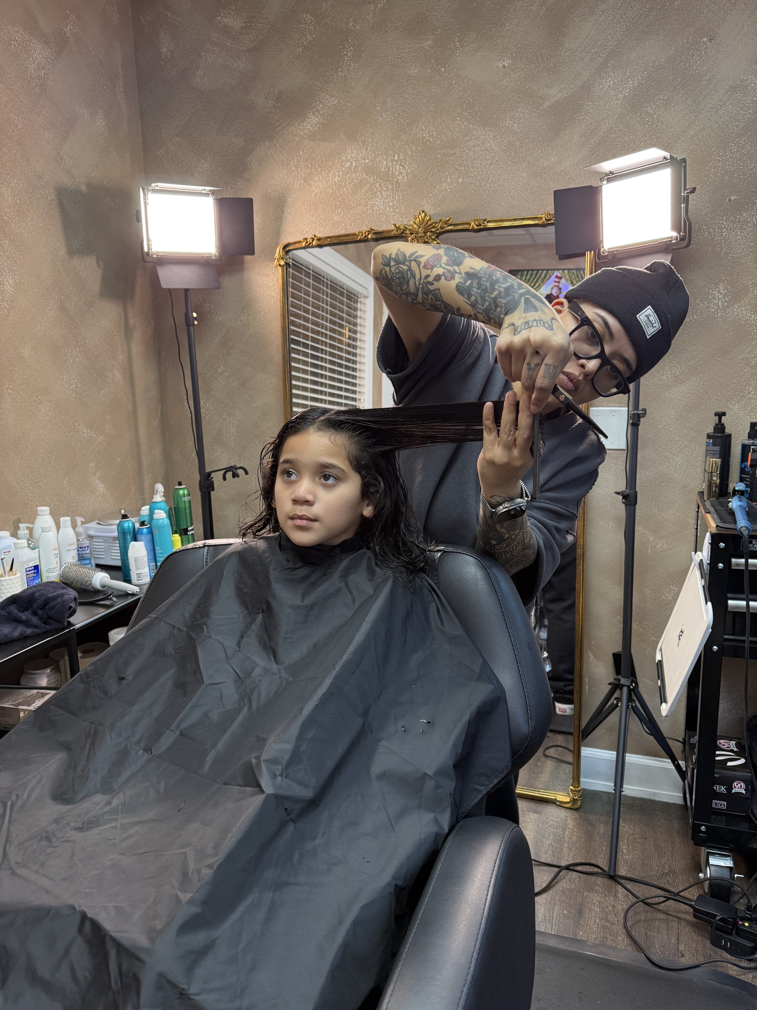A hairstylist with tattoos and glasses cuts a young girl's hair in a salon with professional lighting, hair products, and a mirror.