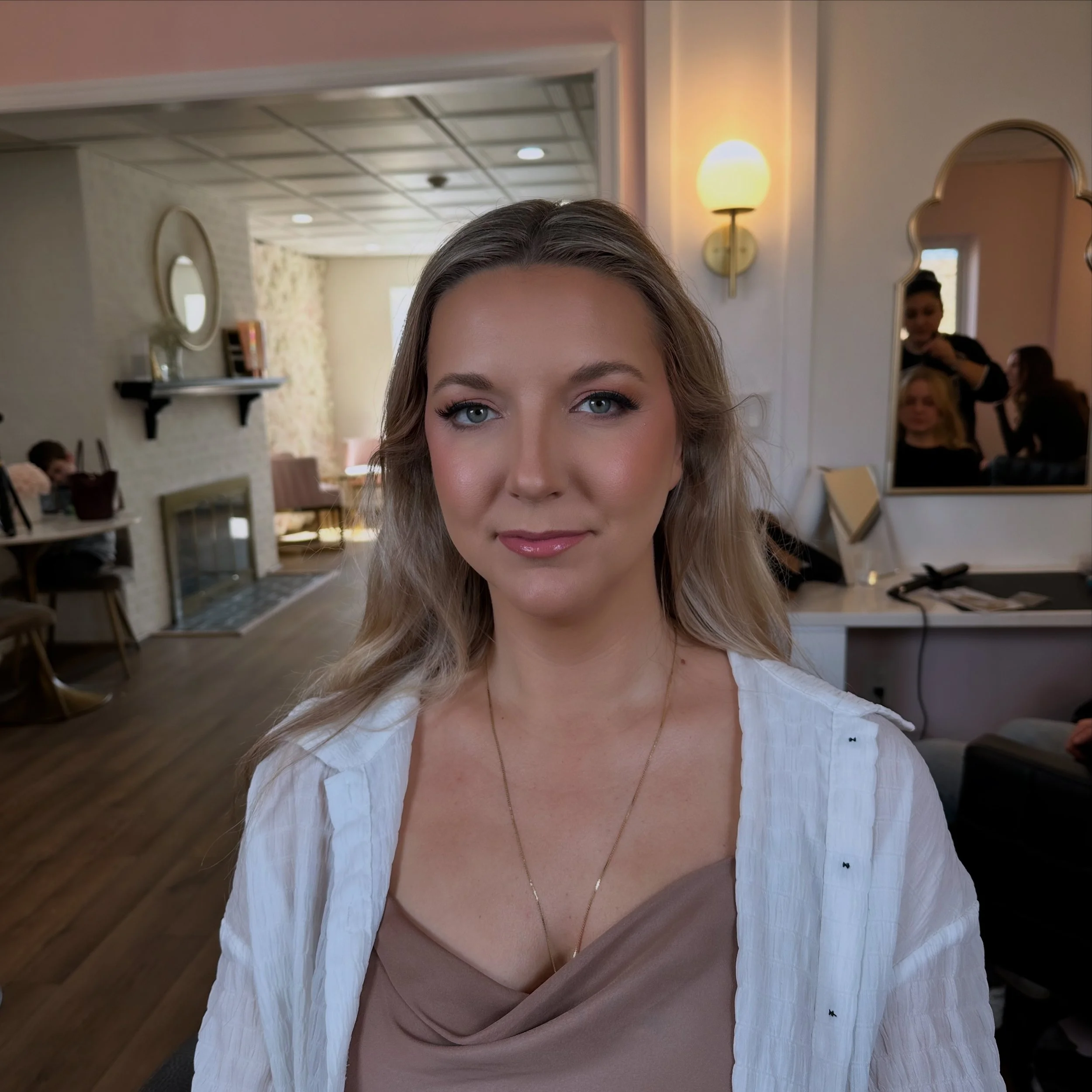 A woman with blonde hair and blue eyes, wearing a white cardigan and beige top, sitting in a room with warm lighting, a fireplace, and a mirror in the background.