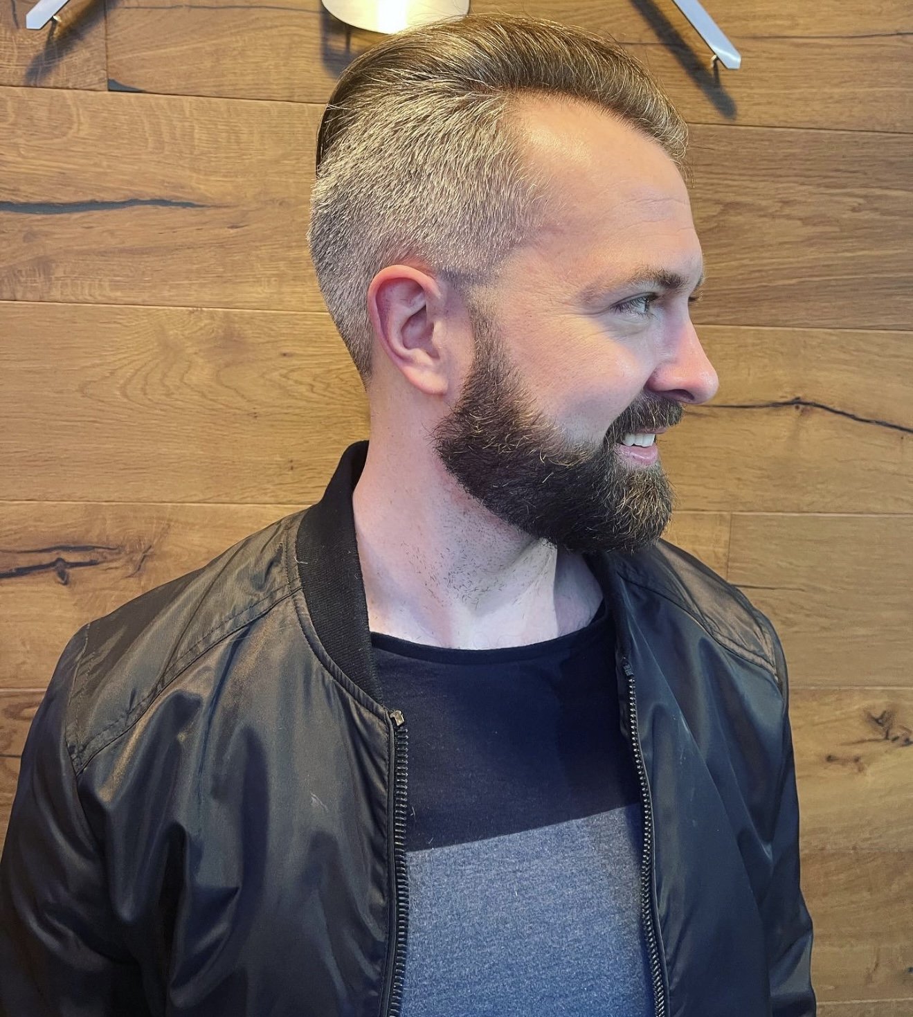 A man with a dark beard and slicked back hair, wearing a black jacket over a gray shirt, smiling and looking to the right, standing against a wooden wall background.