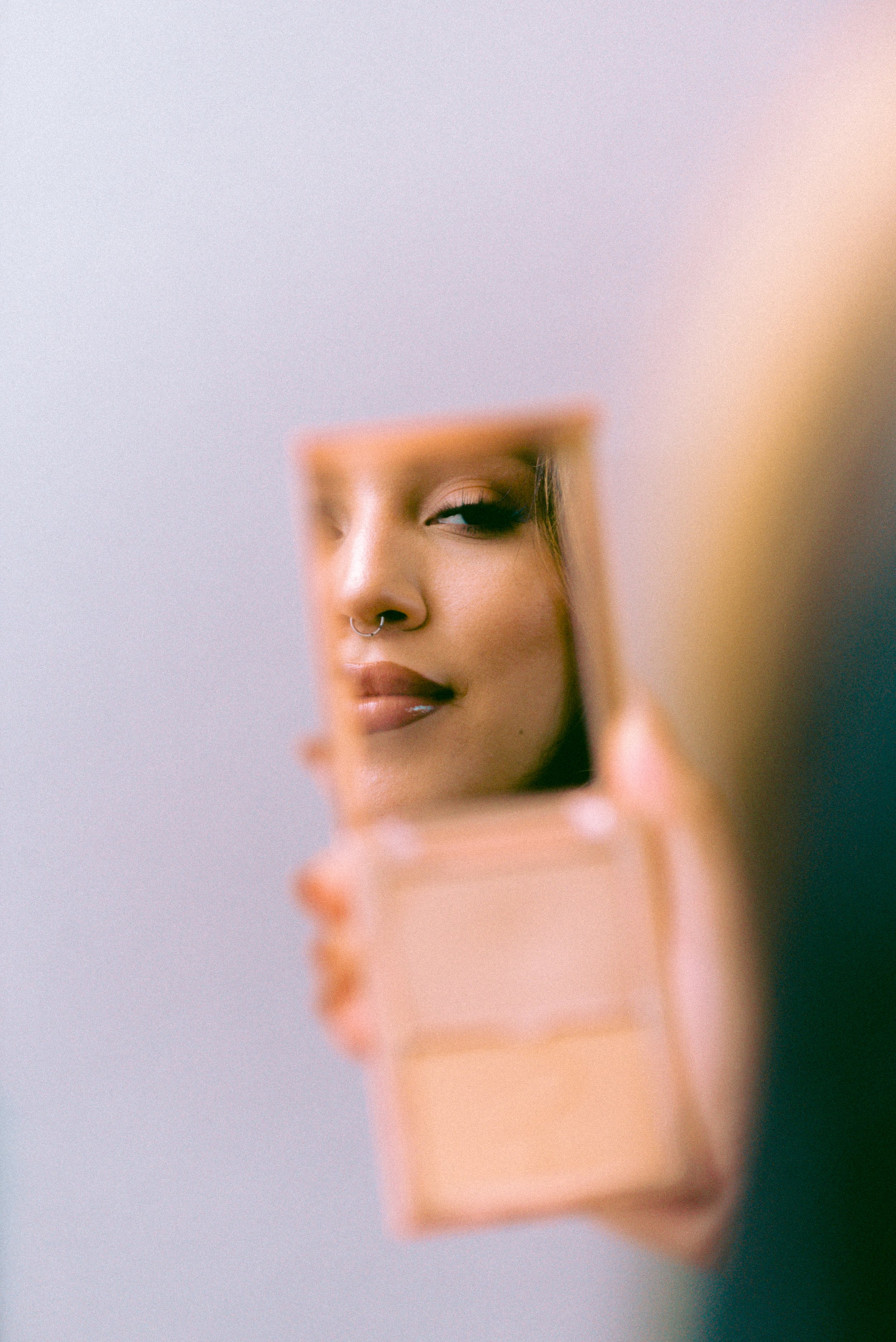 A person holding a small mirror reflecting their face, showing a woman with makeup, a nose ring, and half-closed eyes.