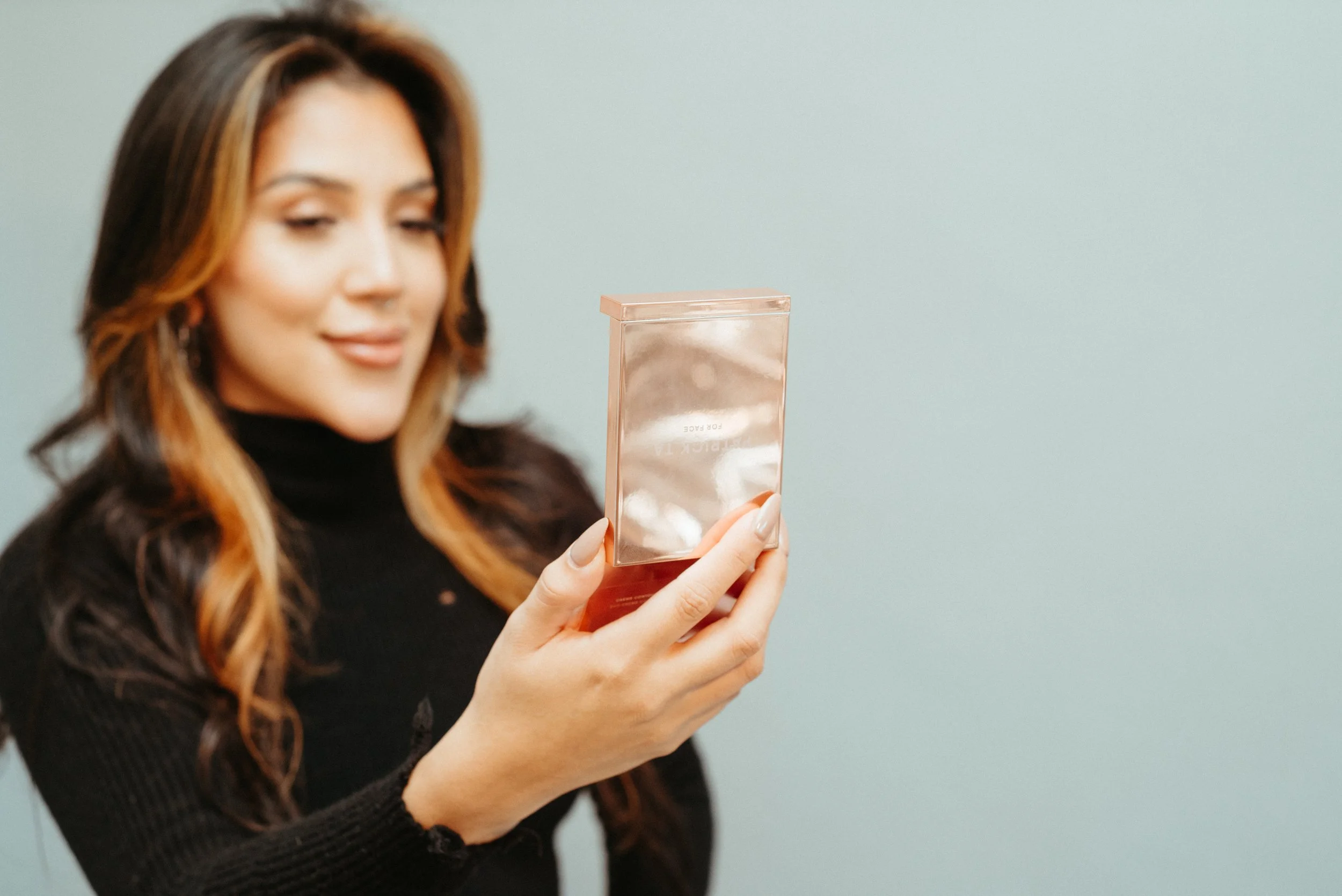 A woman with brown hair in loose waves, wearing a black turtleneck, is holding a clear and pink reflective box close to her face and looking at it with a slight smile.