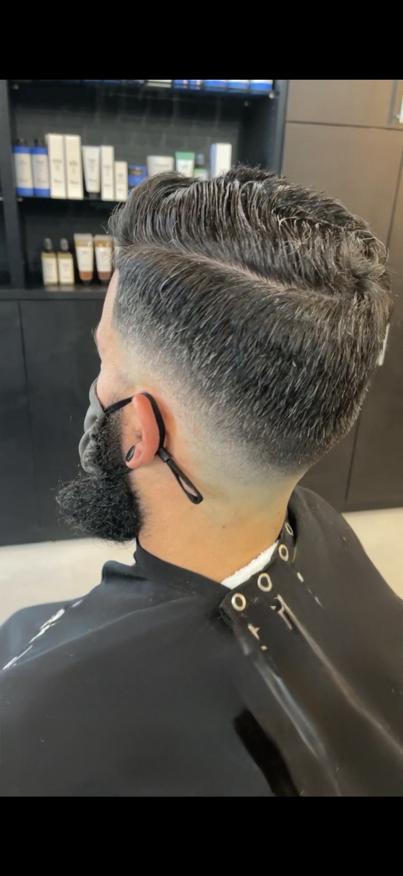 A man with a freshly cut, layered hairstyle with a fade on the sides, wearing a black face mask and a barber cape in a salon.