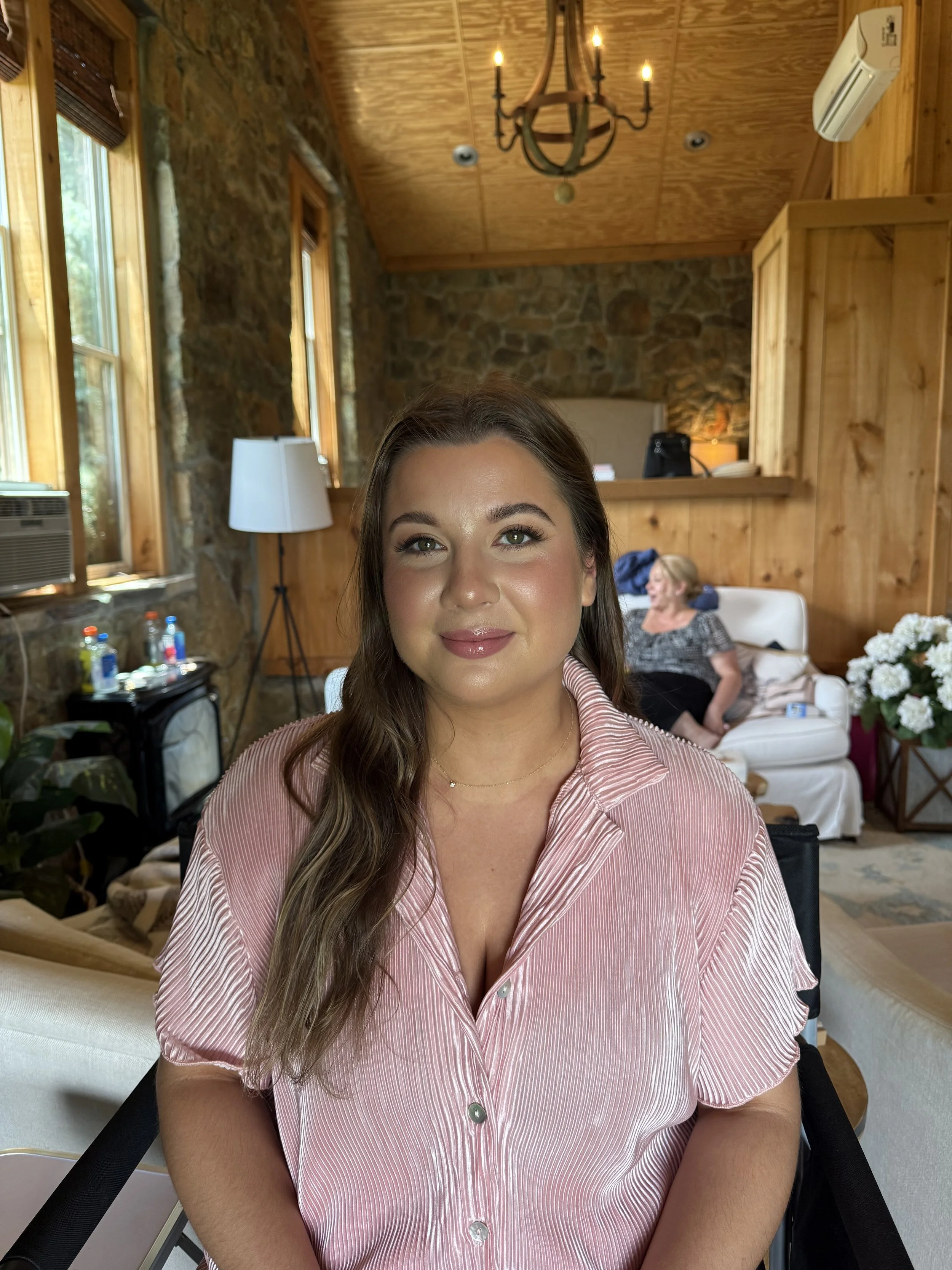 A young woman with long brown hair, wearing a pink striped shirt, sitting indoors with a woman resting on a white couch in the background, stone and wood walls, large windows, and a chandelier.