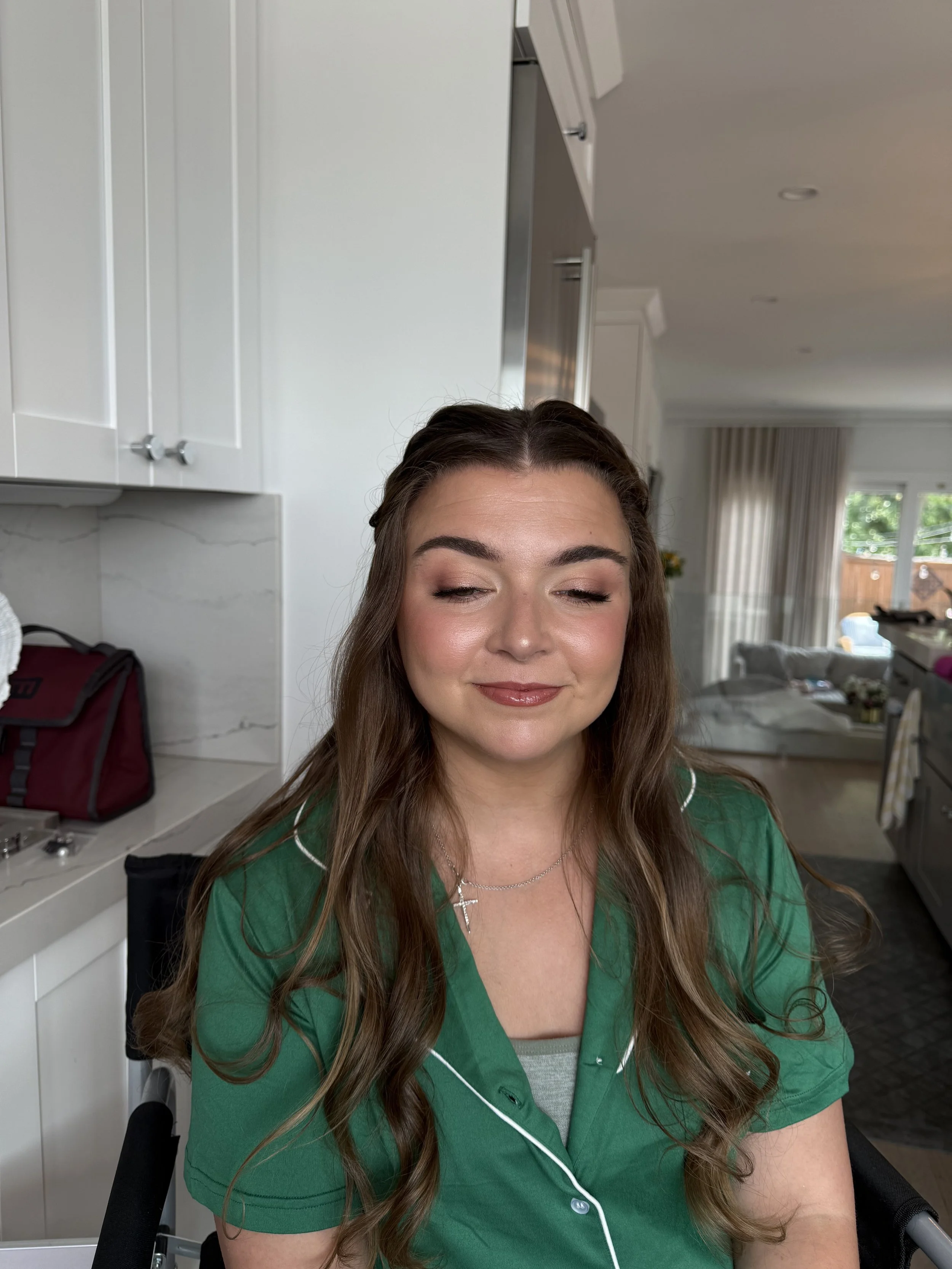 A woman with long wavy brown hair and light makeup in a green pajama top with white piping, sitting in a kitchen with closed eyes and a slight smile.