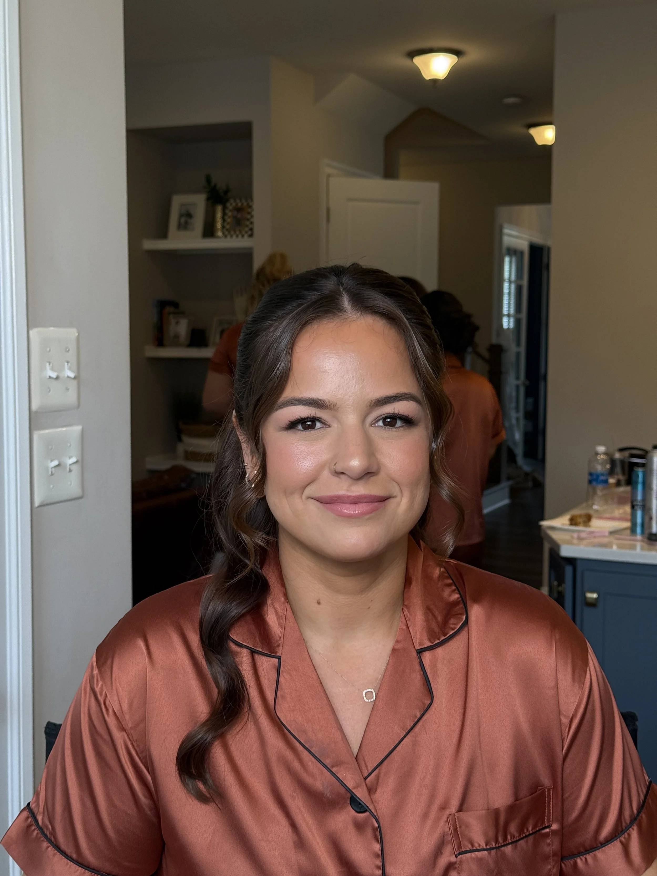 A woman with long brown hair styled in loose waves, wearing a satin rust-colored silk pajama top with black piping, smiling at the camera inside a home.