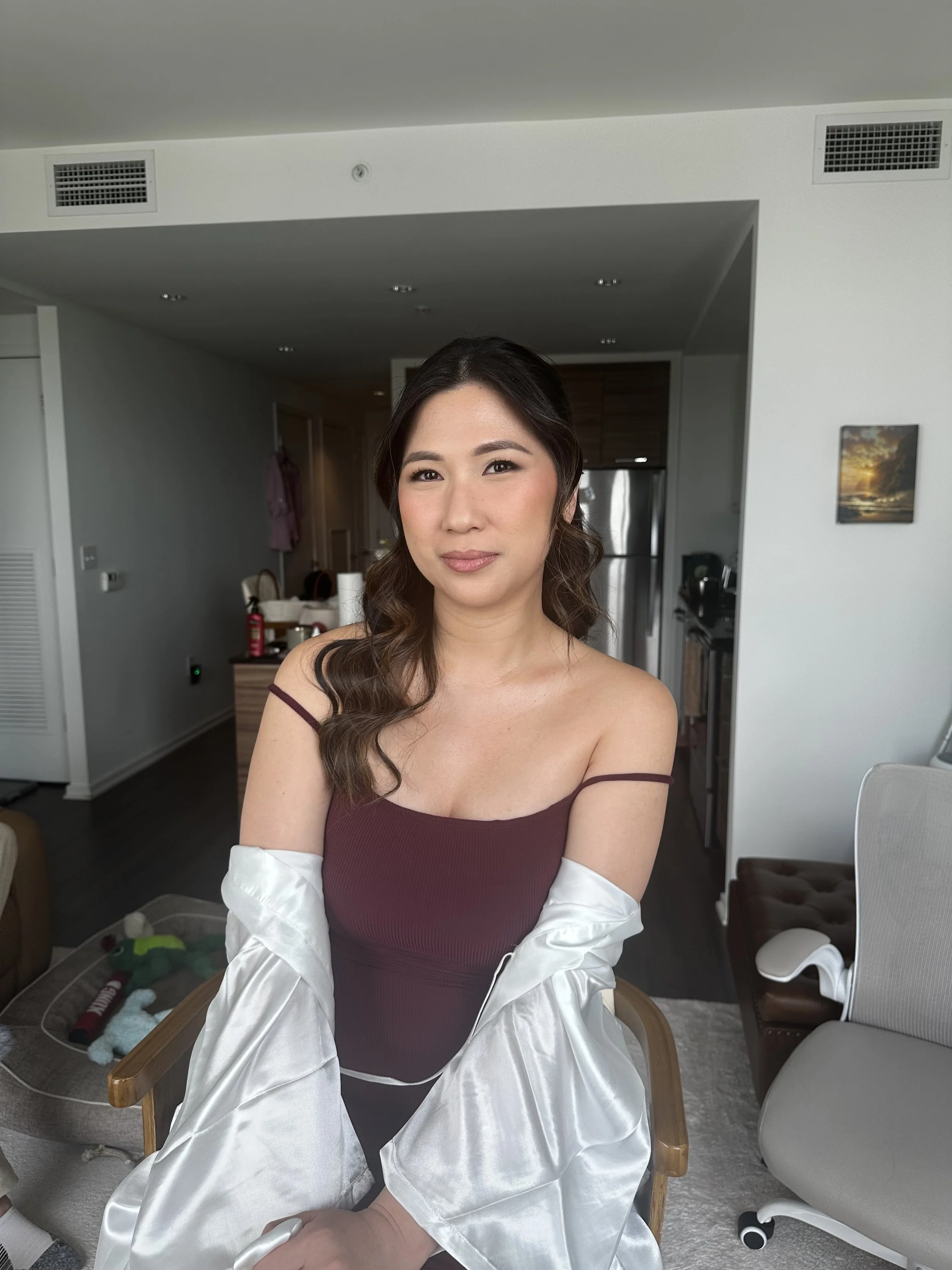 A woman with wavy brown hair and a maroon off-shoulder top sitting on a chair in a modern apartment.