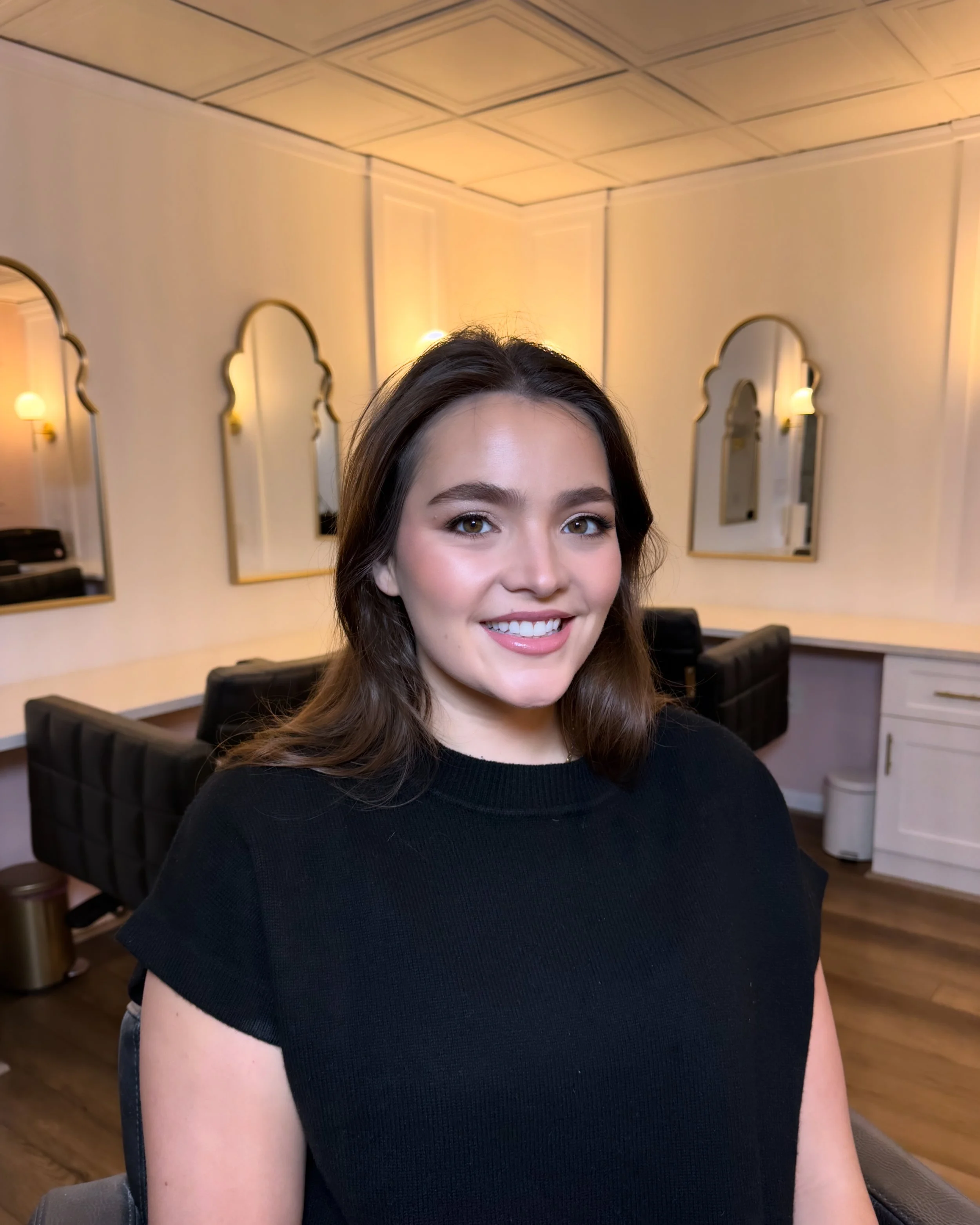 A young woman with brunette hair and a black shirt smiling in a room with mirrors and salon chairs.