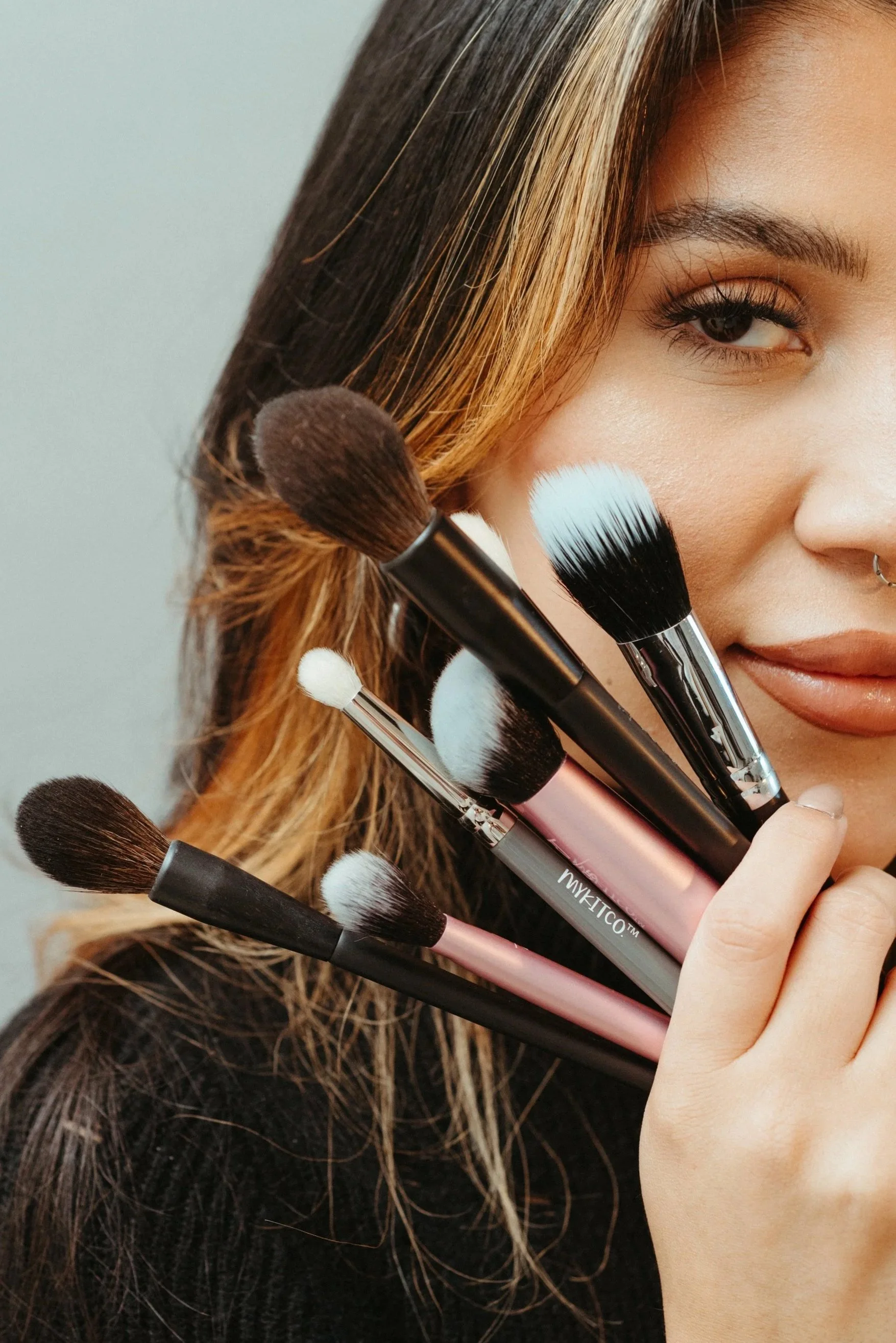 A woman holding various makeup brushes near her face, with visible makeup and hair.