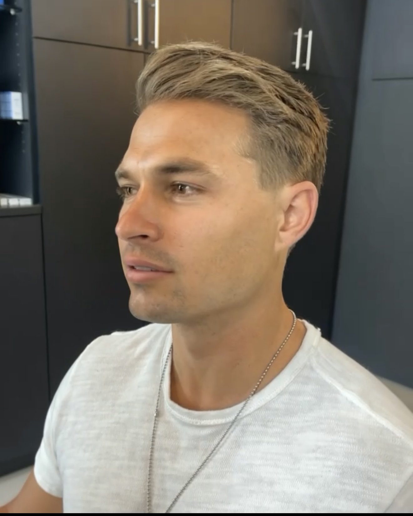 A man with short, neatly styled blonde hair wearing a white t-shirt and a silver chain necklace, sitting indoors near dark-colored cabinets.