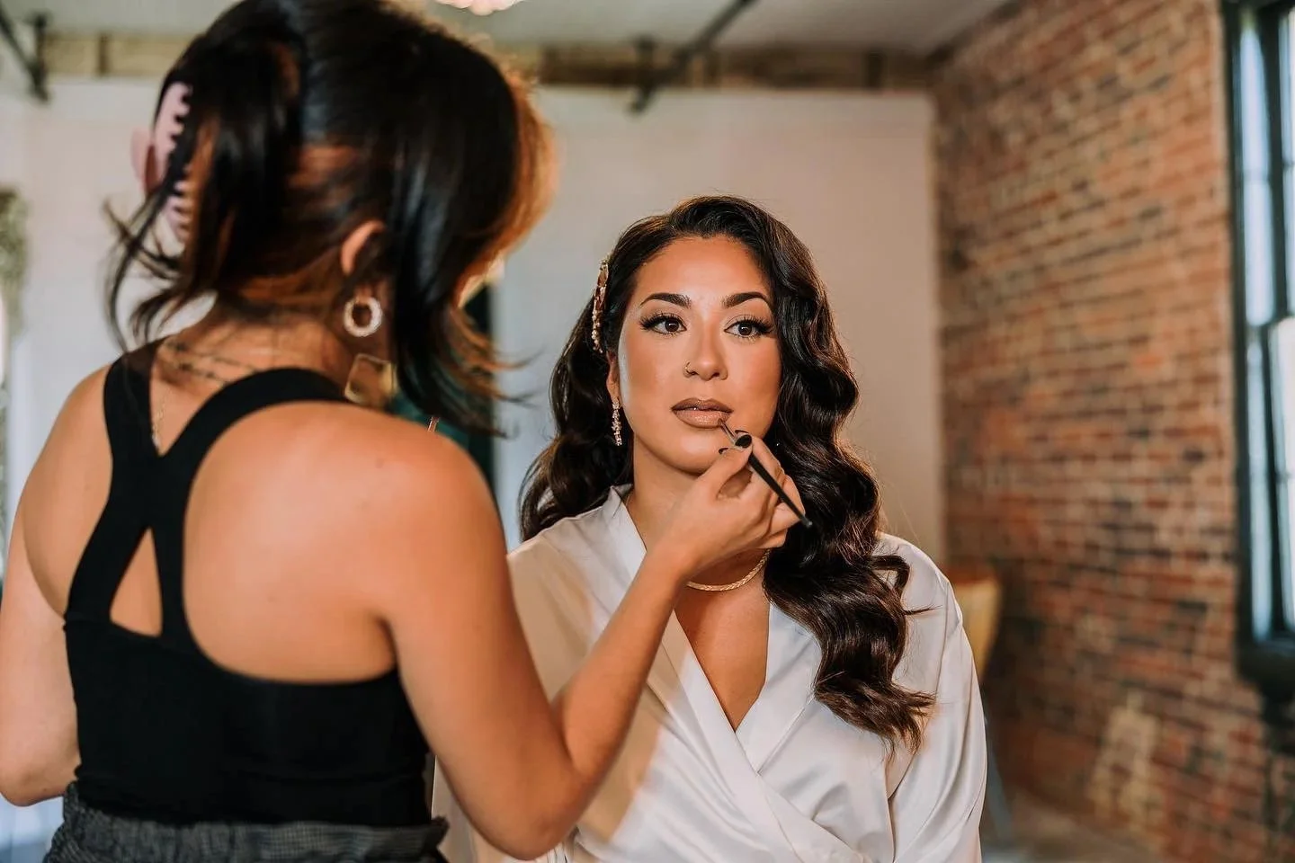 Makeup artist applying lipstick to a woman with wavy dark hair in a white satin robe in a brick-walled room.