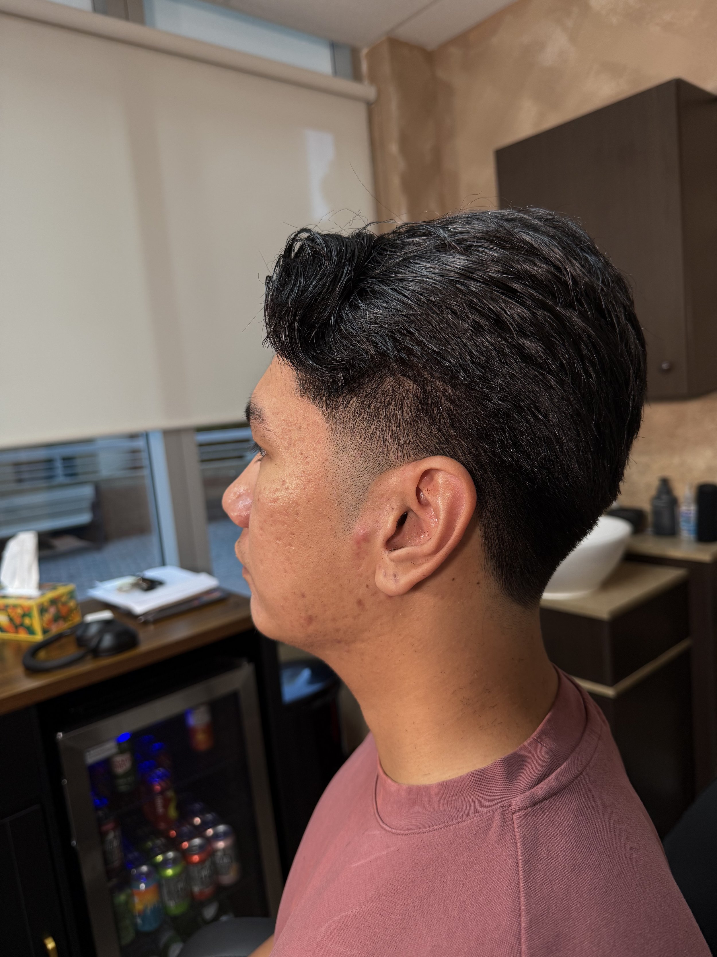 Profile of a young man with a fresh haircut, featuring a fade on the sides and longer, slicked-back hair on top, sitting in a room with a beige wall, a window, and dark furniture behind him.