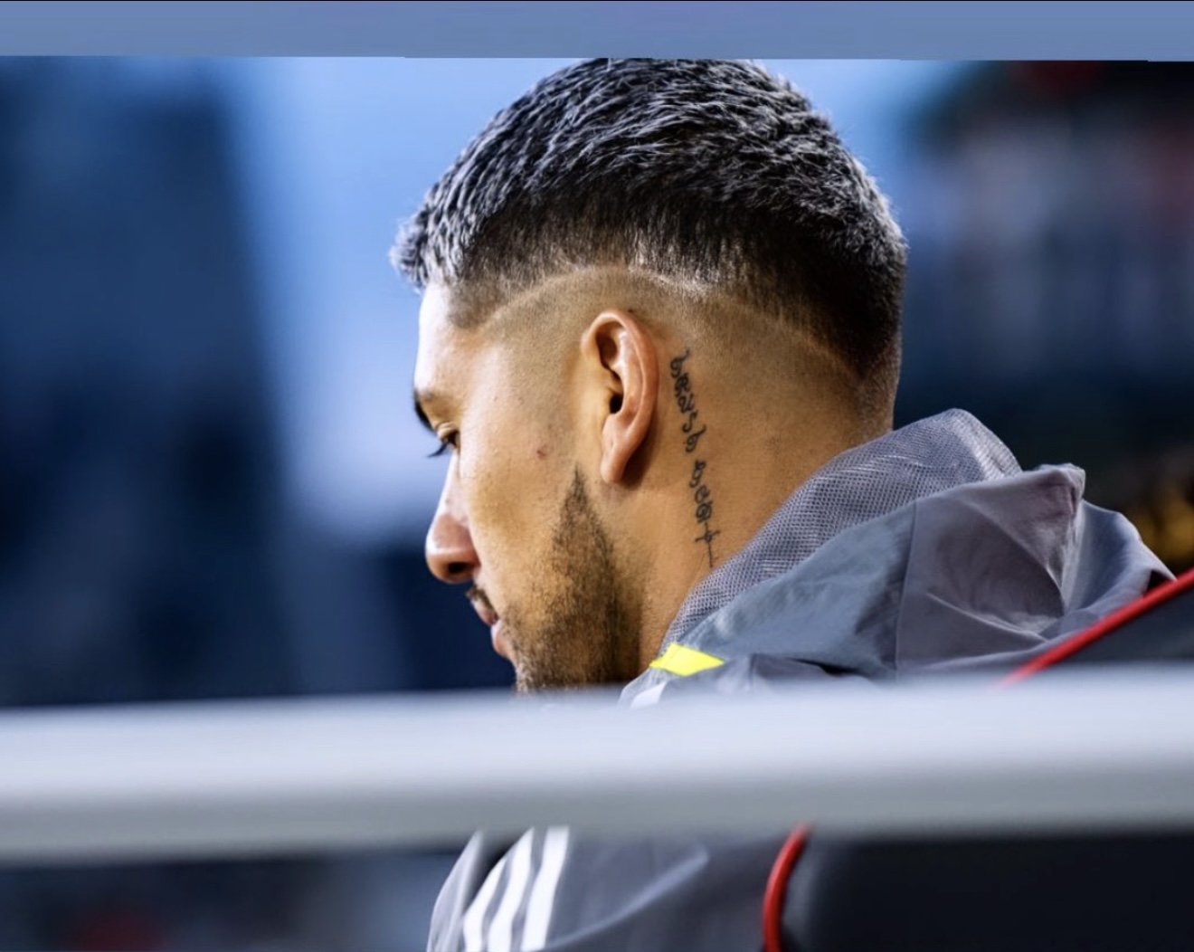Side profile of a man with styled hair and a tattoo on his neck, sitting outdoors wearing a grey hoodie.