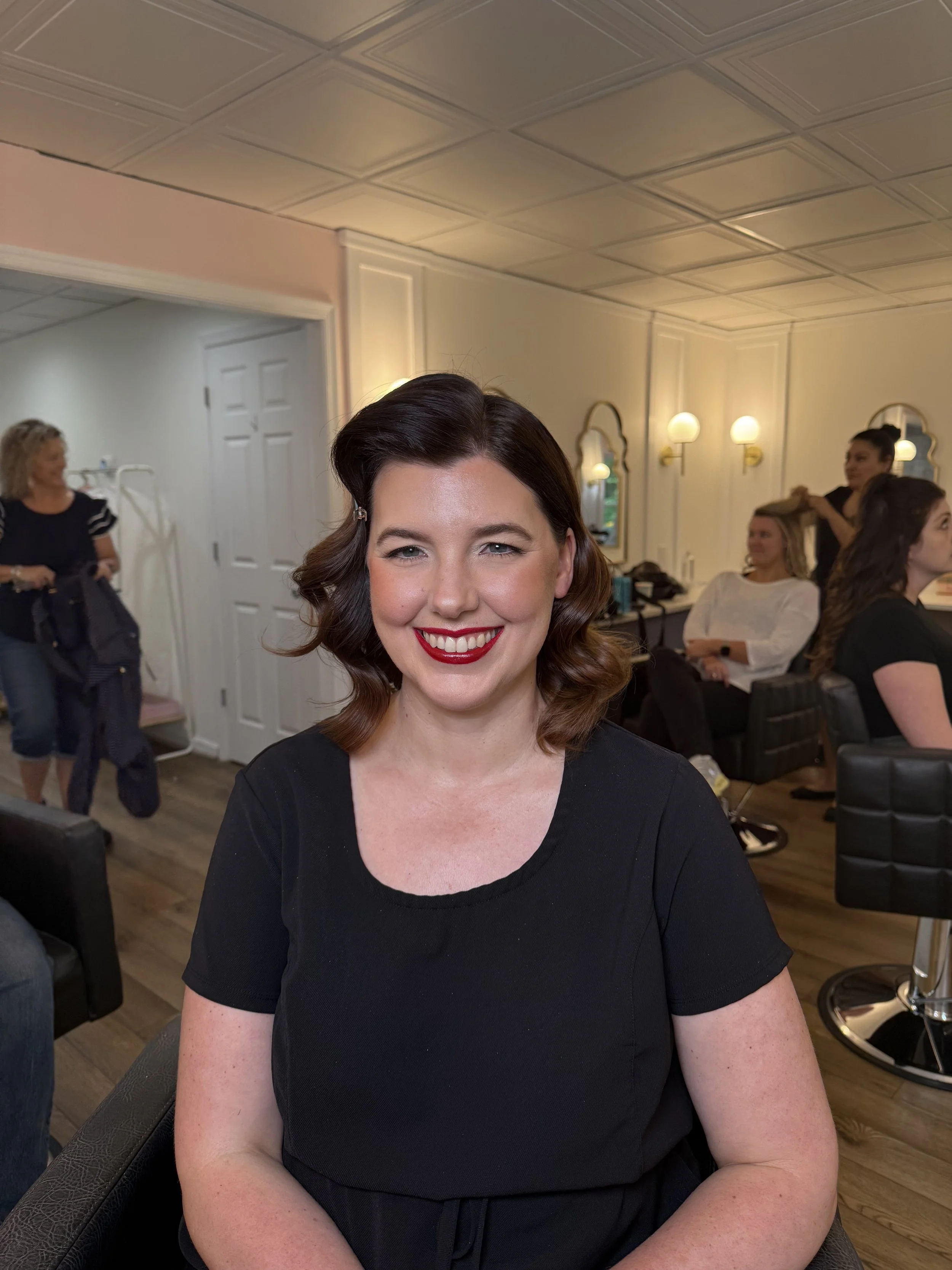 A woman with dark brown hair styled in vintage waves, wearing red lipstick and a black top, smiling at the camera inside a beauty salon.