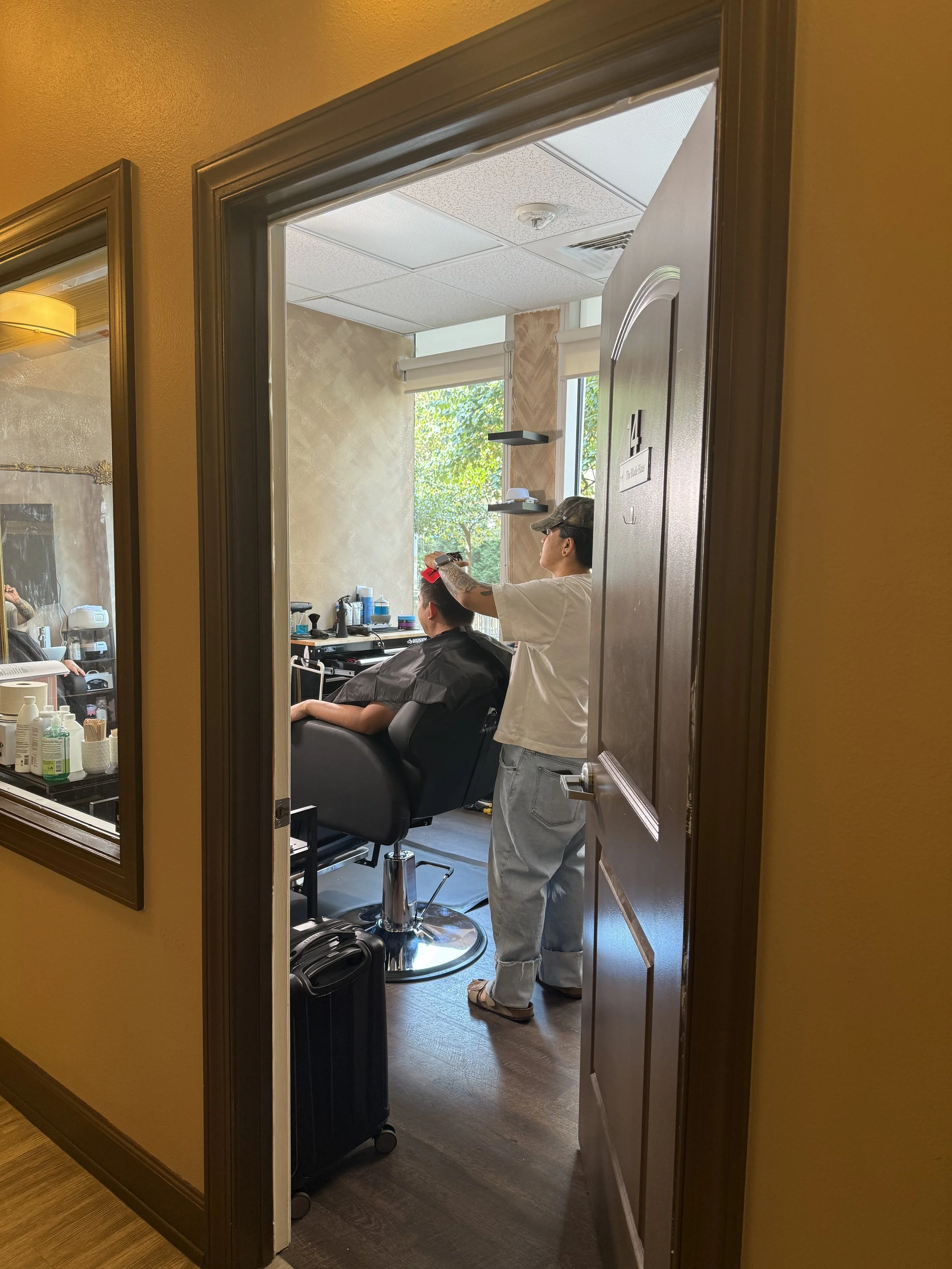 View into a salon room where a stylist is applying hair color or treatment to a client seated in a salon chair. The stylist is standing, wearing a white shirt, and the client is covered with a black cape. The room has a large window with sunlight and
