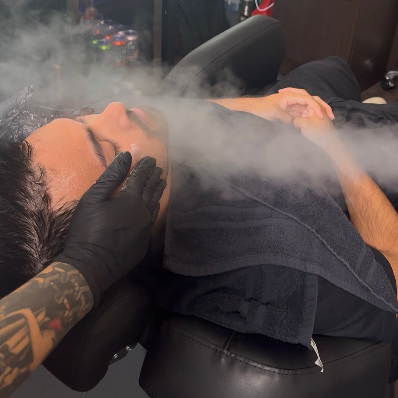 Person receiving a facial treatment with steam, lying on a black chair, while a practitioner with black gloves and tattoos gently touches their face.