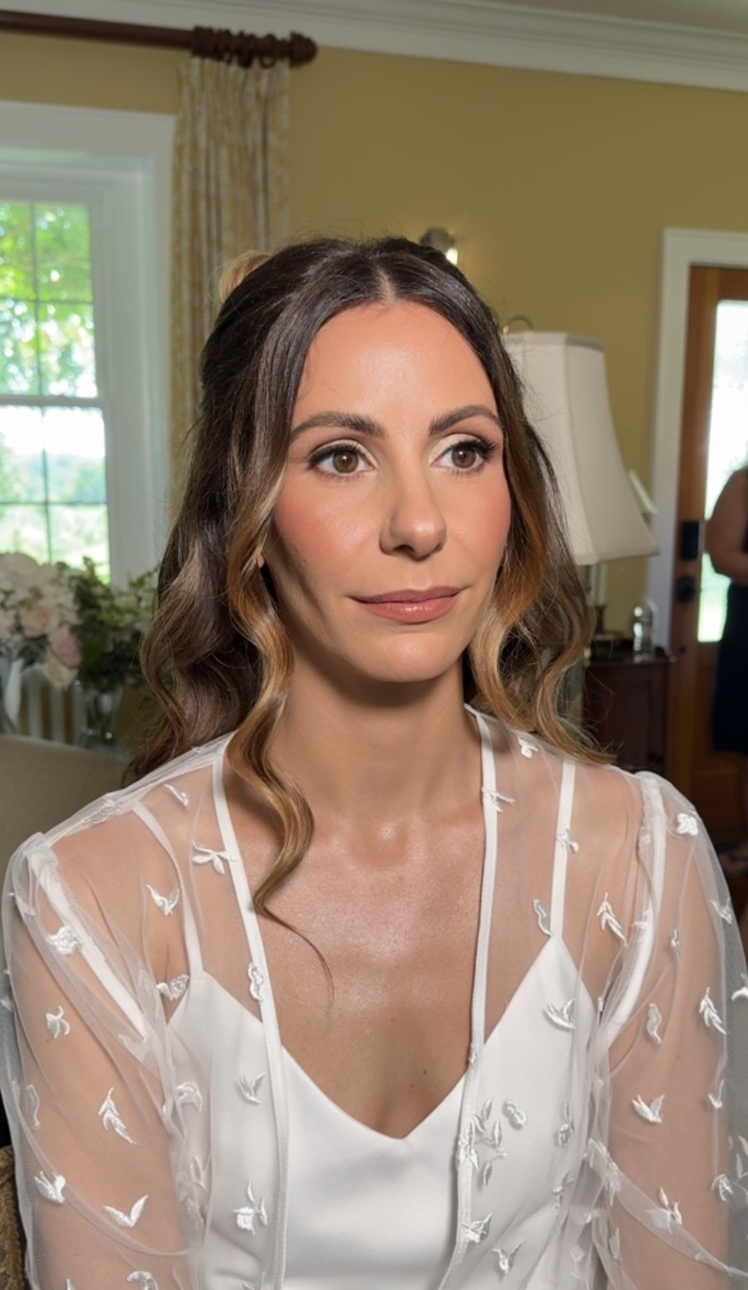 Close-up of a woman with wavy brown hair and a white dress with sheer sleeves and embroidery, sitting in a well-lit room with a window and floral arrangement in the background.