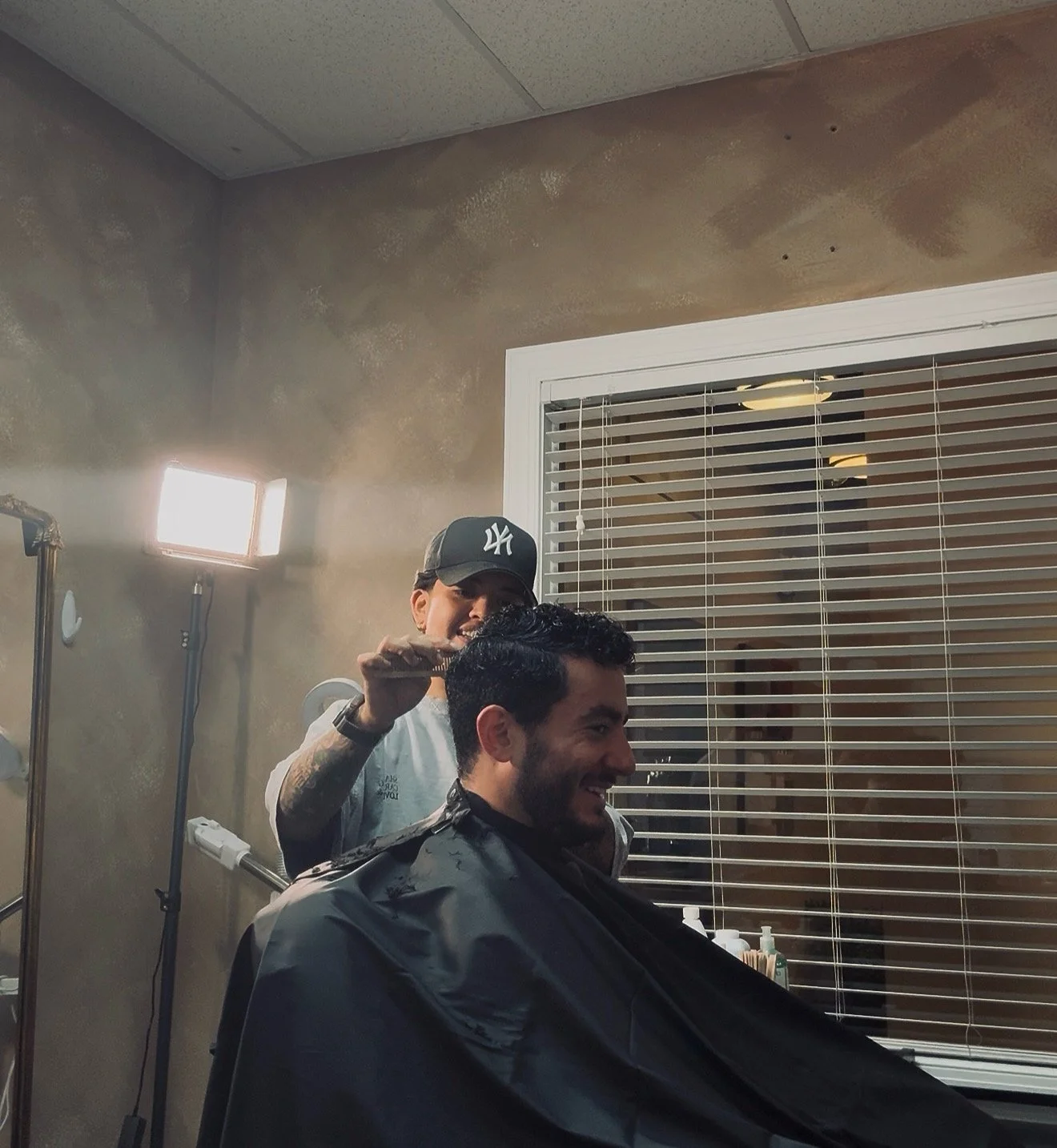 A man with dark hair and beard getting a haircut by a barber wearing a dark NY Yankees cap in a barber shop, with a large window with blinds behind them and bright light on the left.