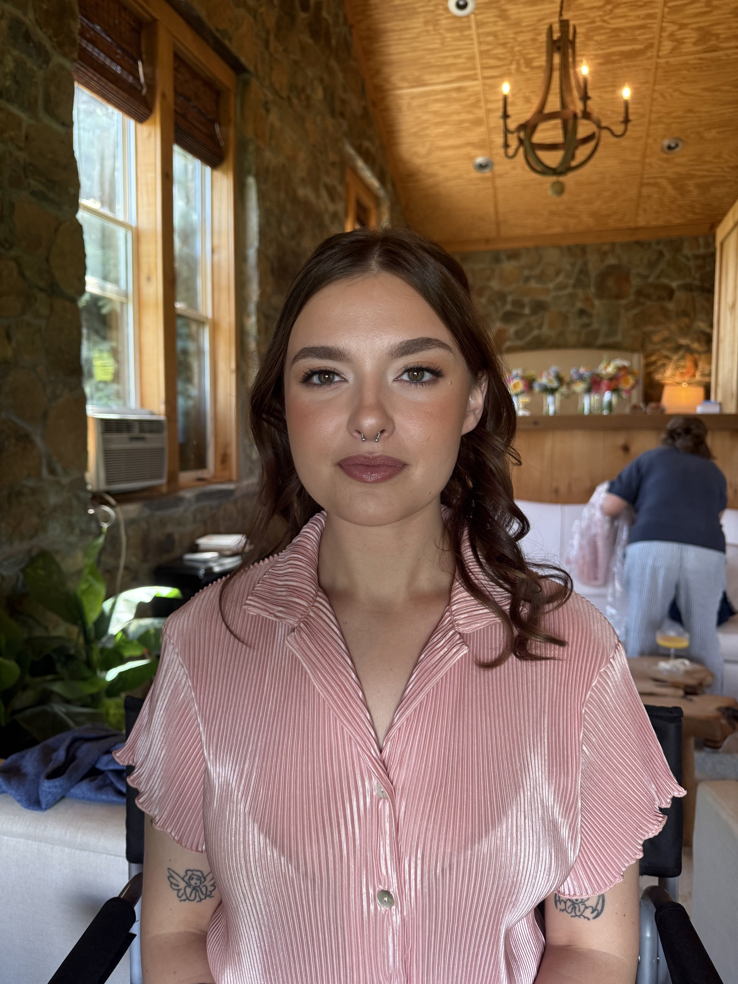 A young woman with shoulder-length brown hair, wearing a pink and white striped blouse, sitting indoors with wooden walls and ceiling, candle chandelier, and a background of a woman arranging flowers.