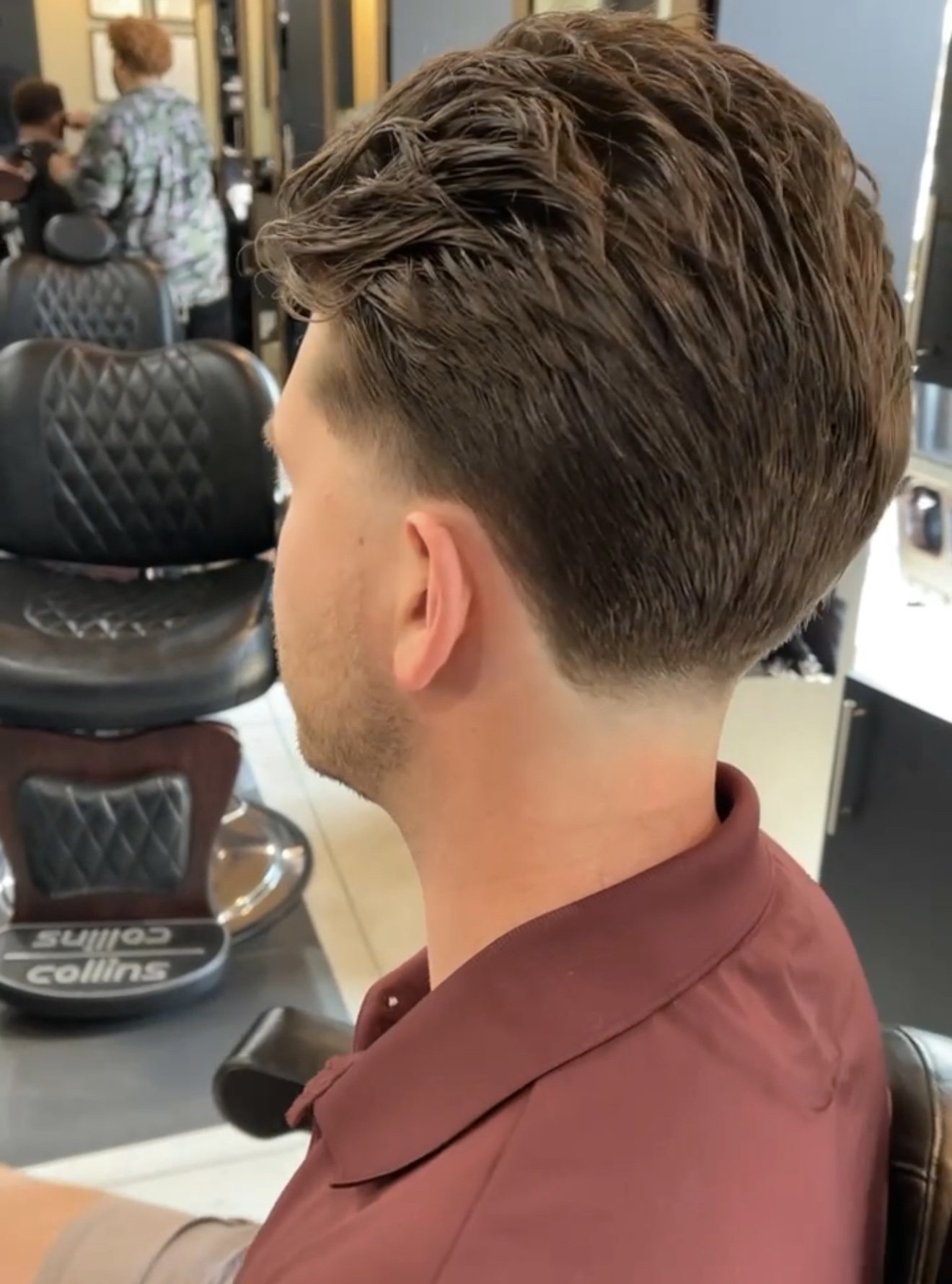 Side view of a man with a fresh, textured undercut hairstyle, sitting in a barbershop. The background shows other clients and barbershop chairs.