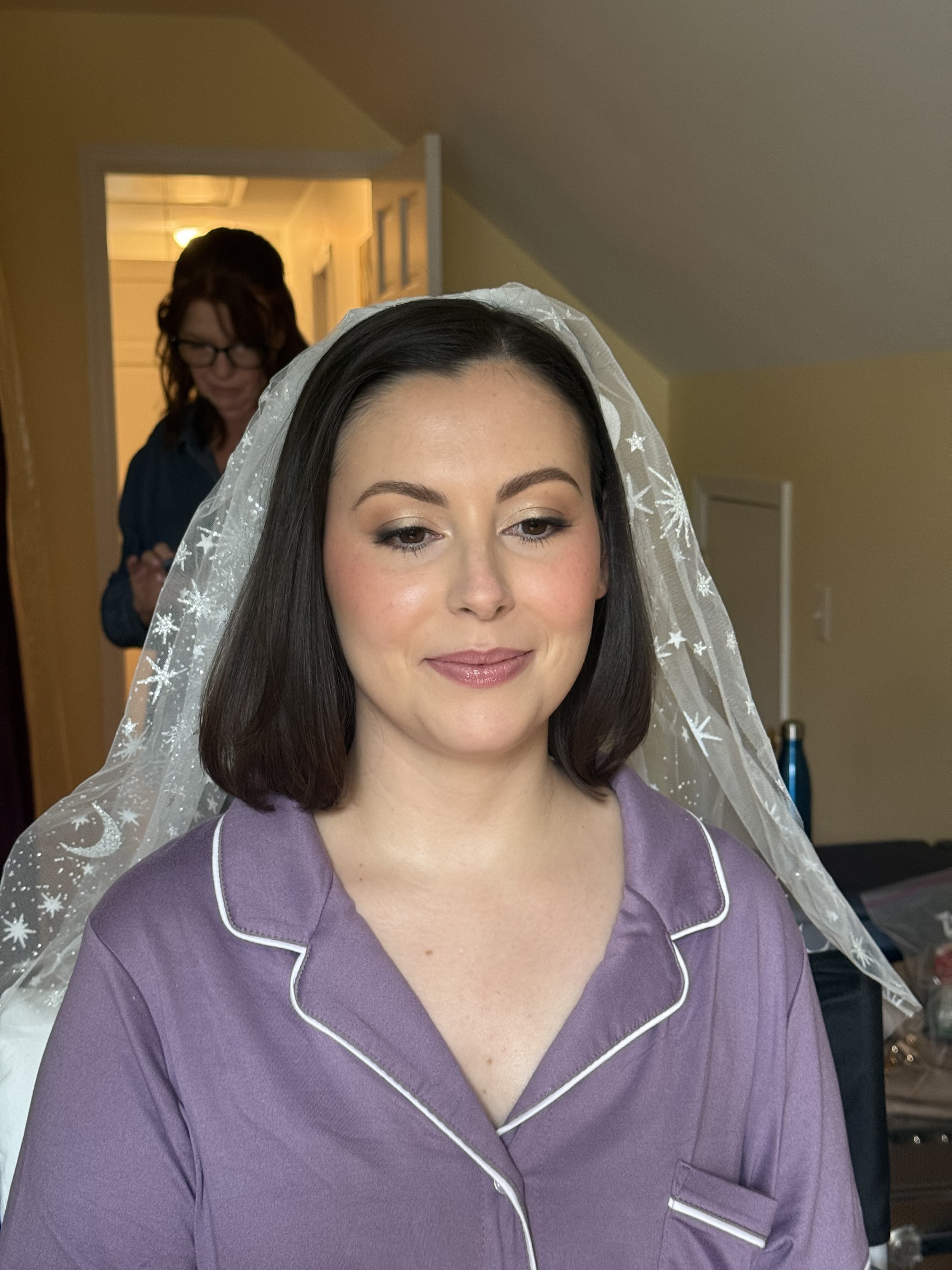 A woman with shoulder-length dark hair, wearing a purple pajama top with white piping and a sheer veil with snowflake and star patterns behind her, smiling softly. In the background, a woman with red hair and glasses is standing in a doorway, blurred