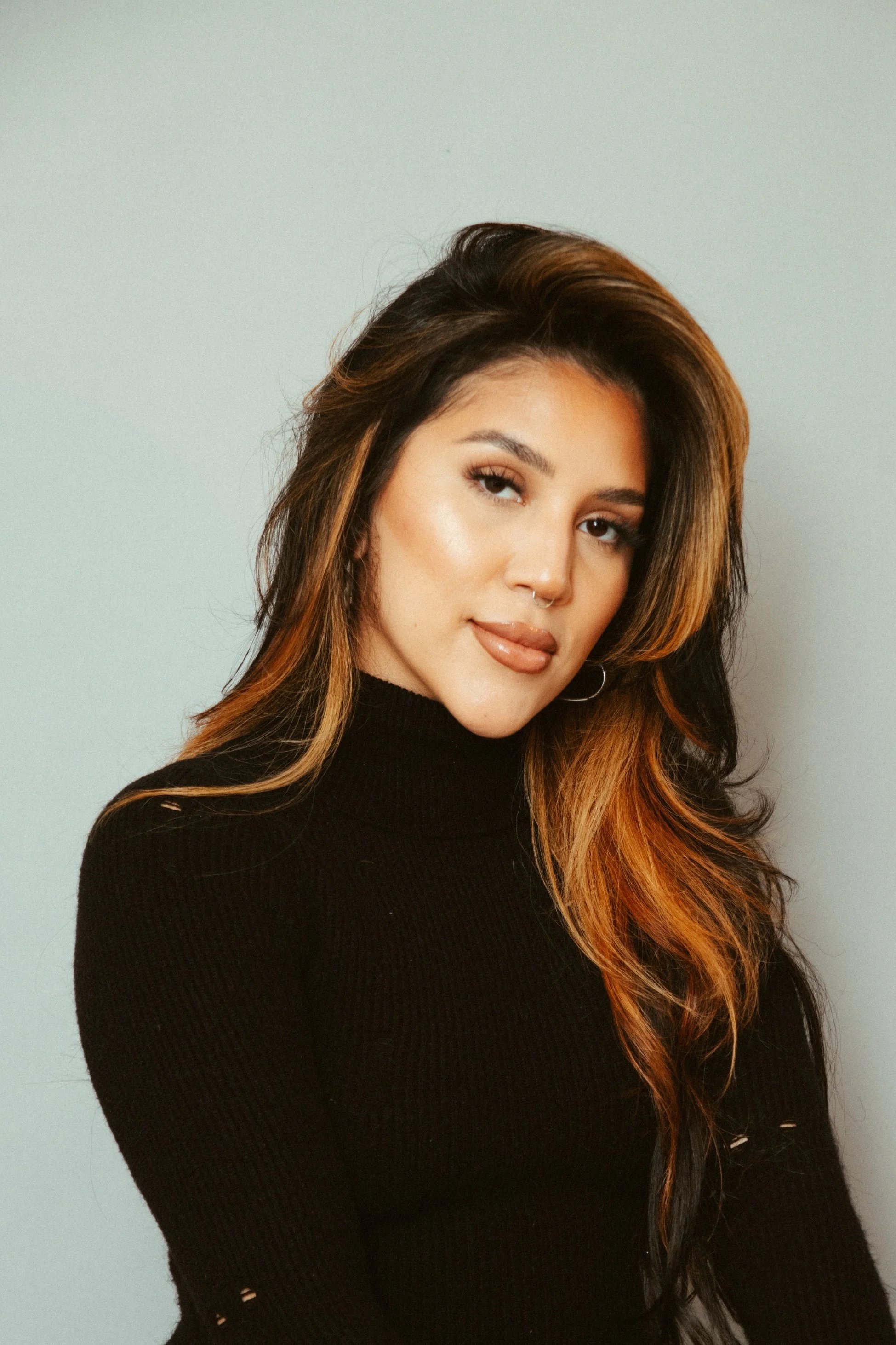 Portrait of a young woman with long, wavy brown hair and light makeup, wearing a black turtleneck sweater, standing against a plain light gray background.