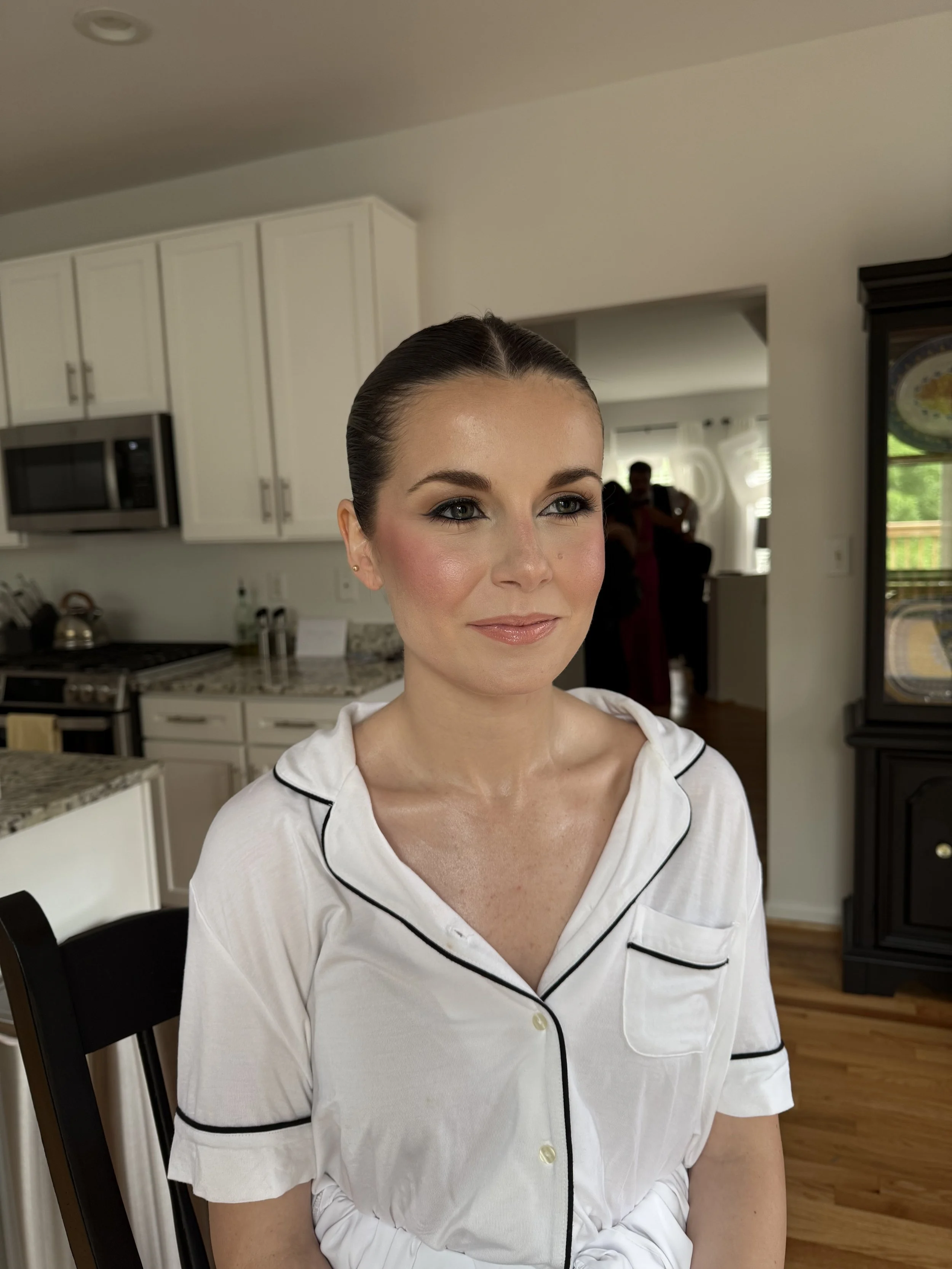 A woman with makeup and hair styled back, wearing a white satin pajama top, sitting indoors in a kitchen.