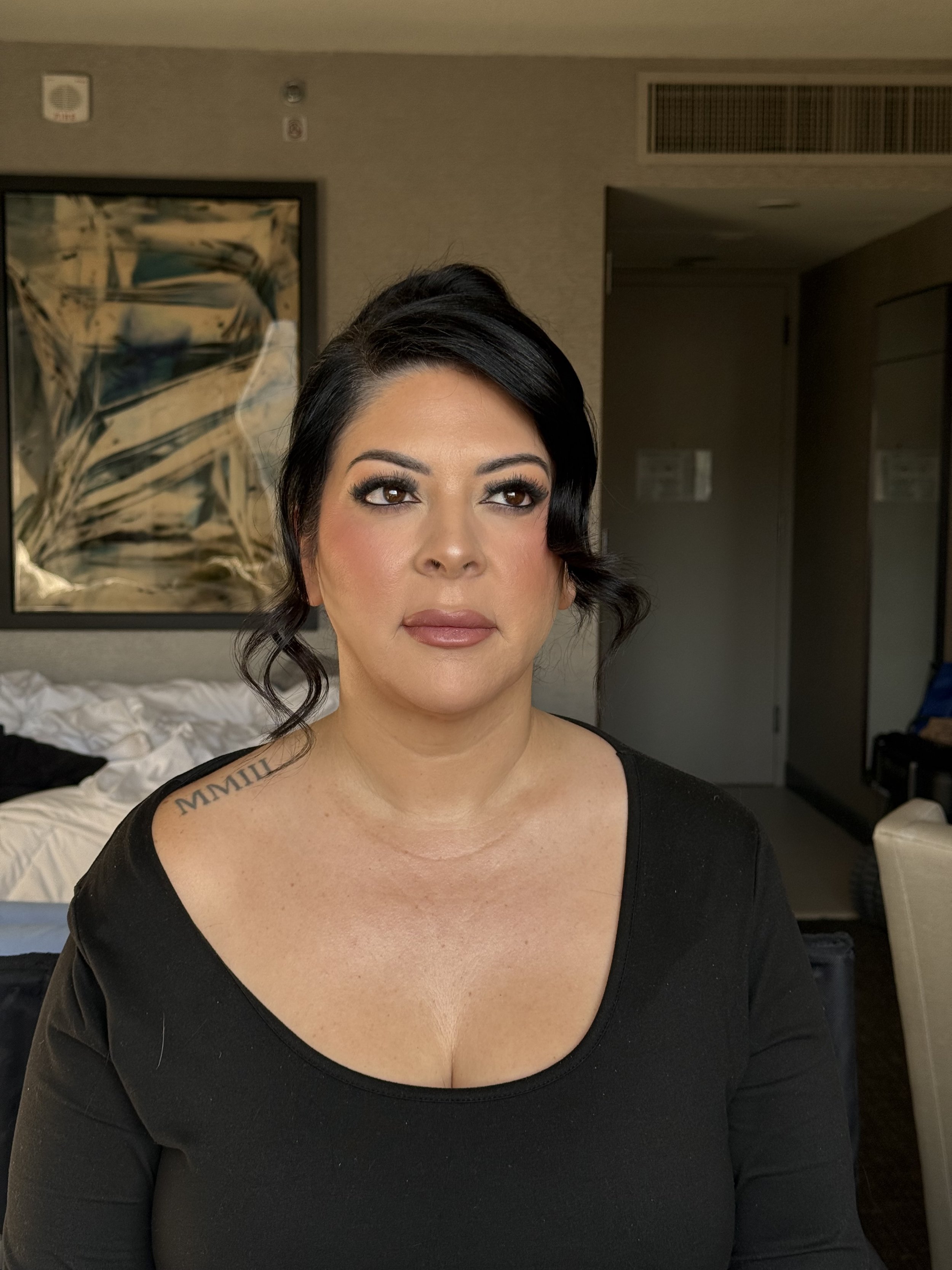 A woman with black hair styled in loose waves, makeup, and a black top sitting in a hotel room or office with abstract art on the wall behind her.