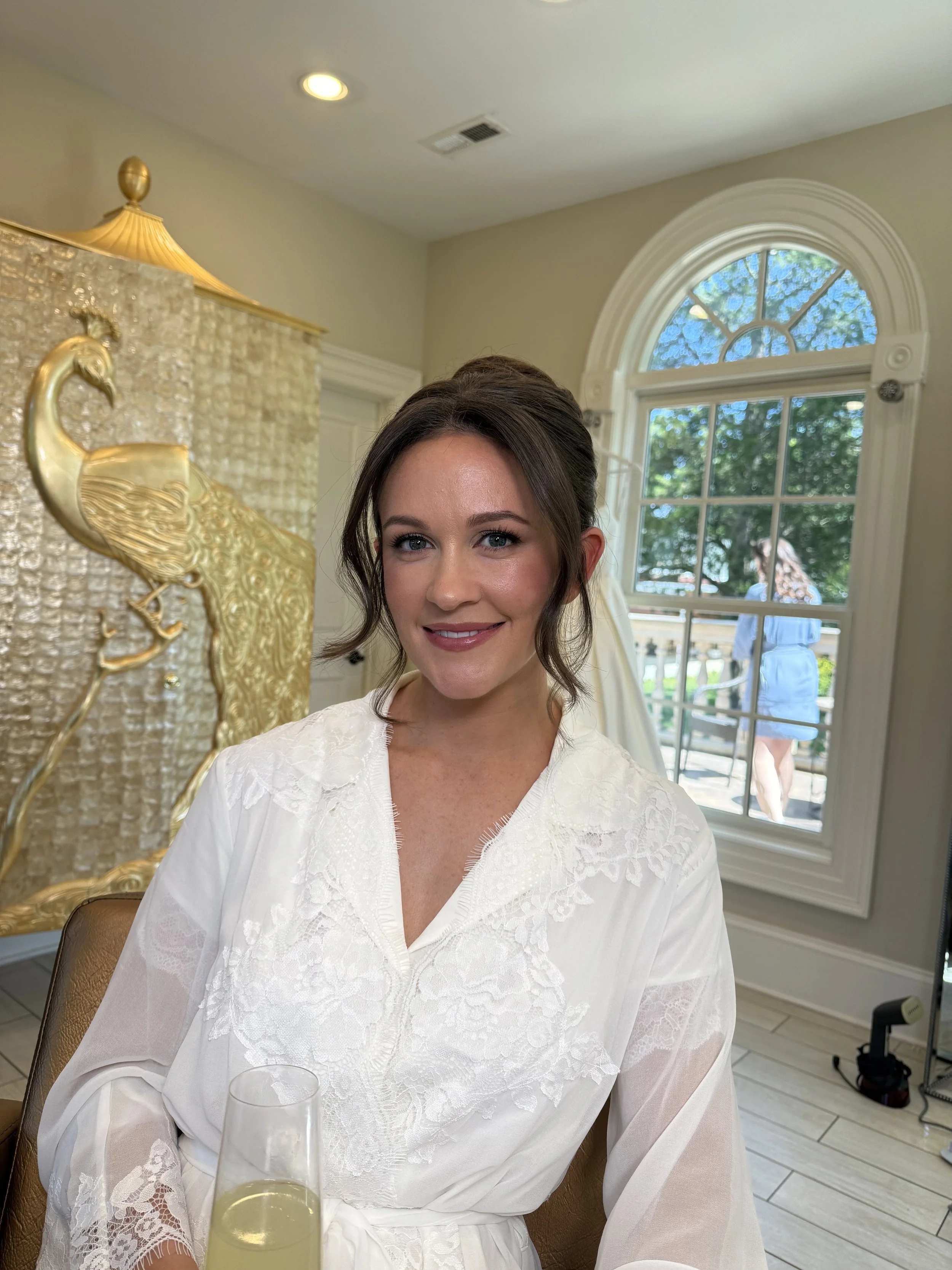 A woman in a white lace dress sitting in a room with a large arched window, decorated with a gold peacock artwork behind her, holding a glass of champagne, smiling at the camera.
