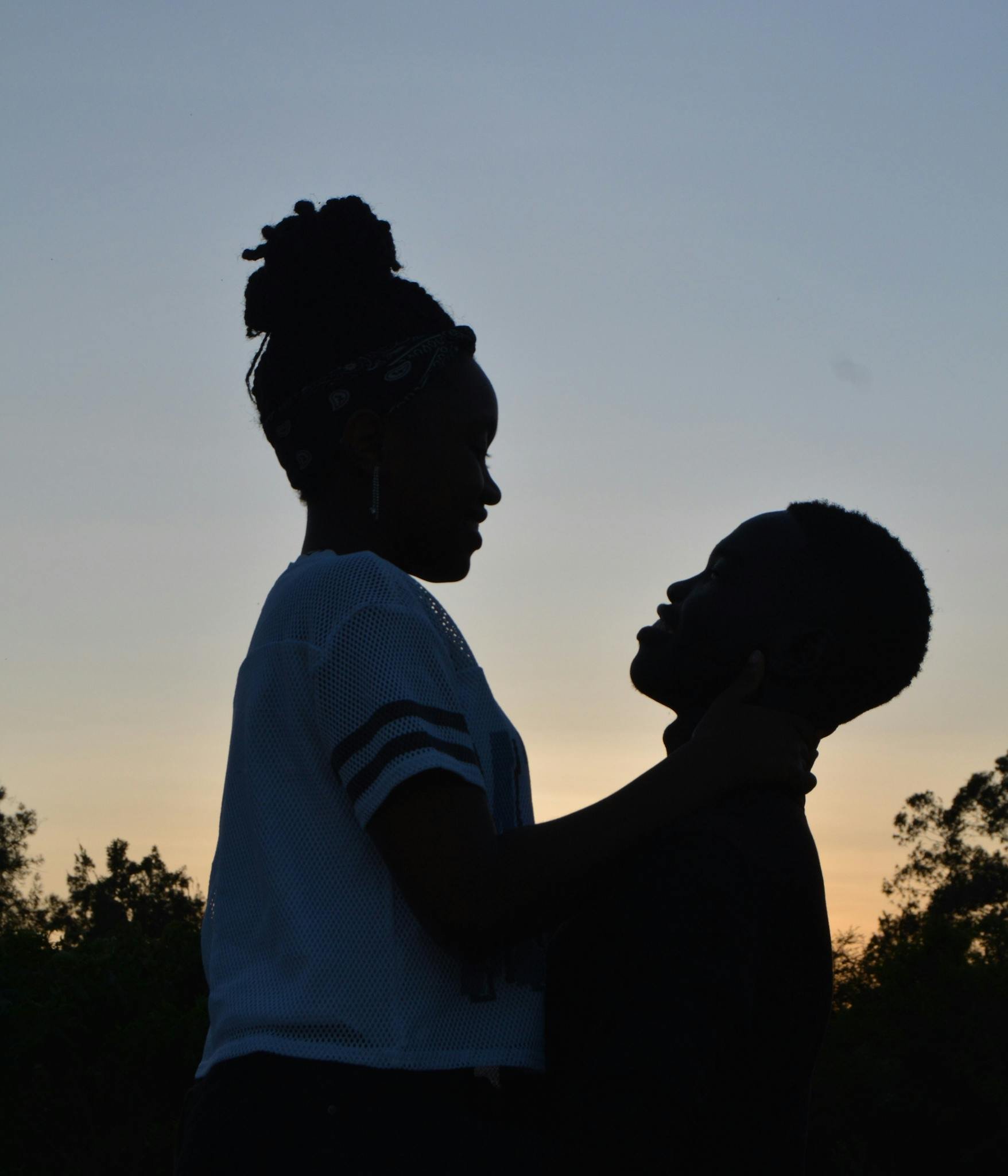 Silhouette of a woman holding a young boy's face against her chest during sunset with trees in the background.