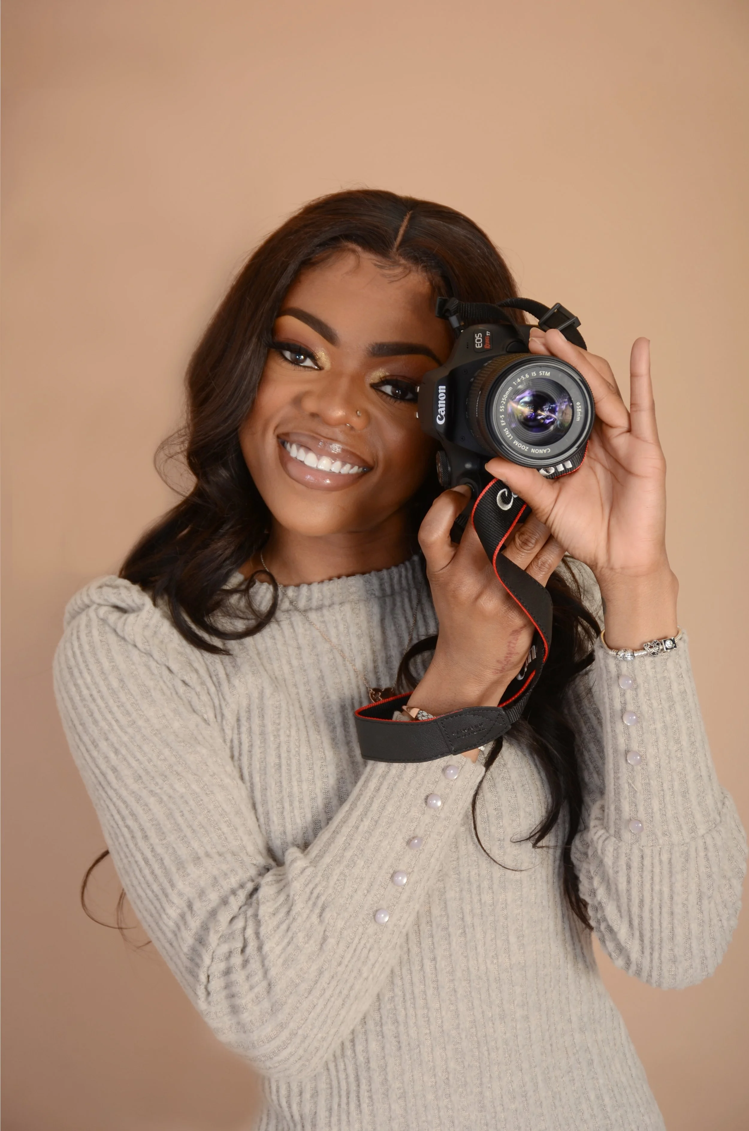 A smiling woman holding a Canon camera close to her face, wearing a beige sweater with pearl buttons and a silver bracelet, with a plain beige background.