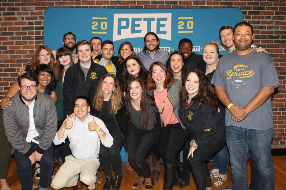 Group of people gathered together at a political event for Pete in 2020, standing in front of a blue backdrop with Pete's name and campaign symbols, smiling and posing for the photo.