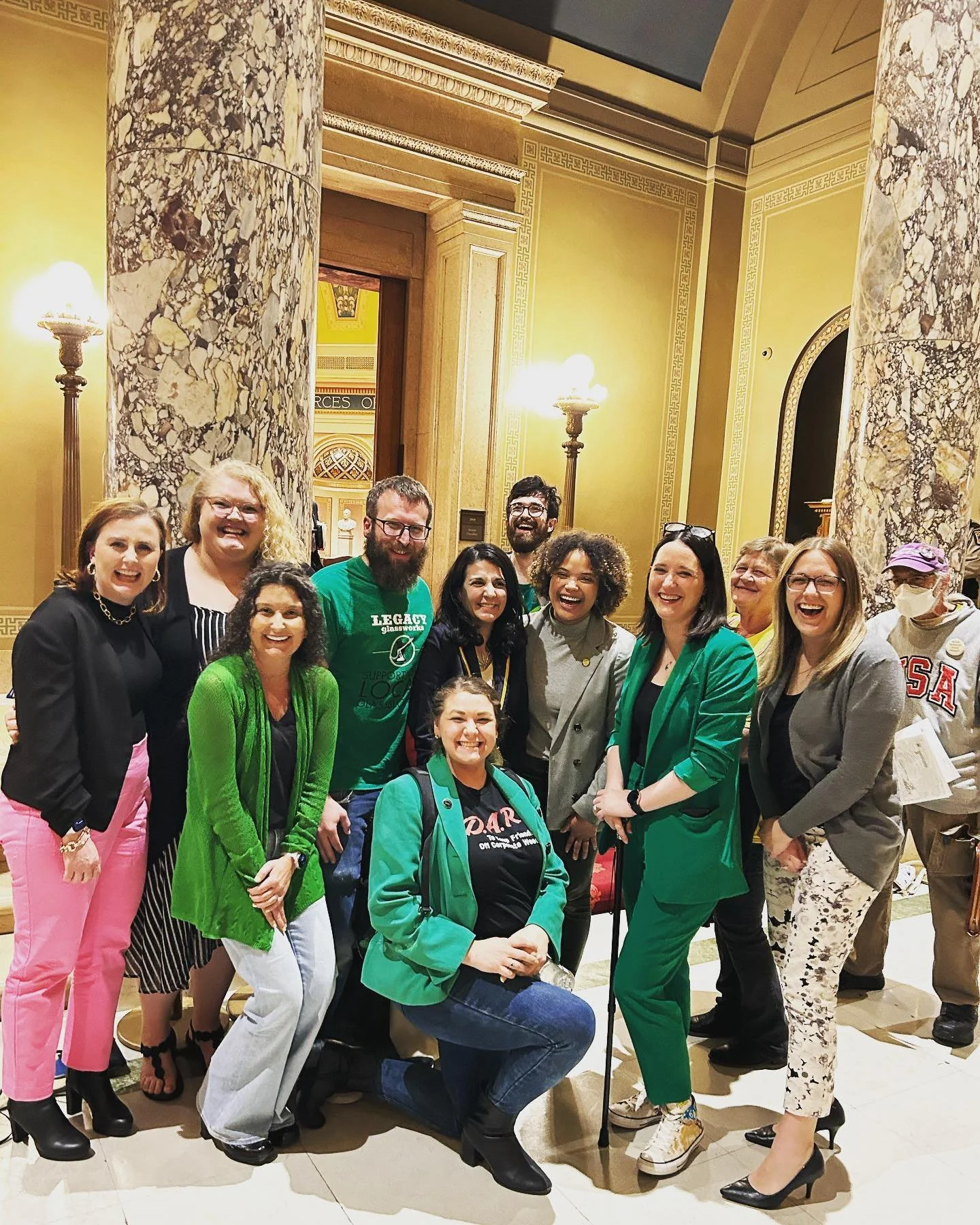 A group of fifteen diverse people smiling and laughing inside a grand, ornately decorated building with marble columns and warm lighting.
