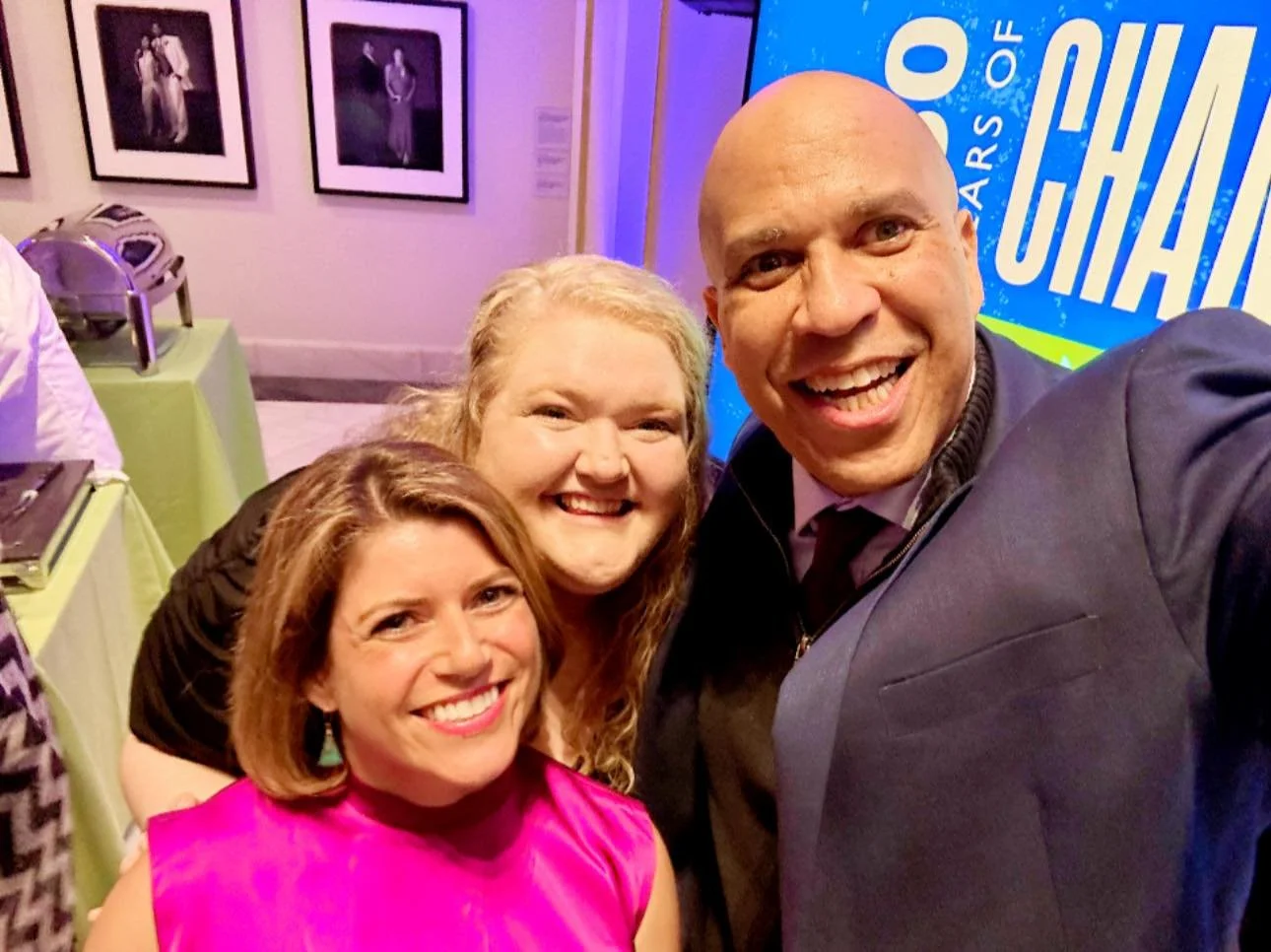 Three smiling people taking a selfie together at an event with framed photographs on the wall and a sign in the background.