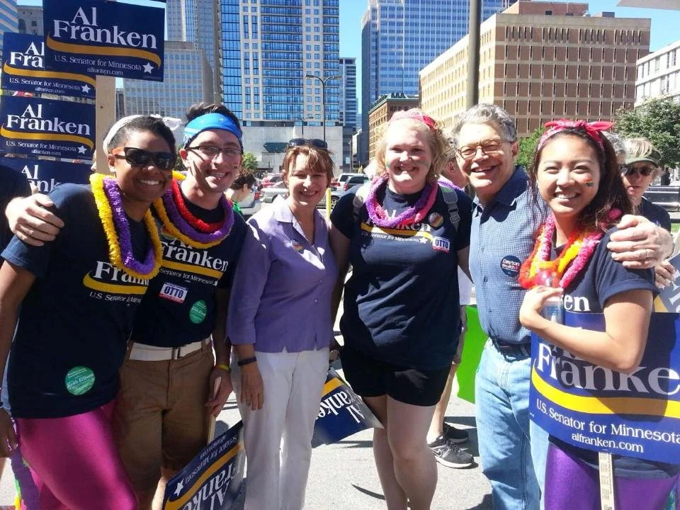 Group of people smiling outdoors during a political rally, holding campaign signs for Al Franken, U.S. Senator for Minnesota, with city tall buildings in the background.