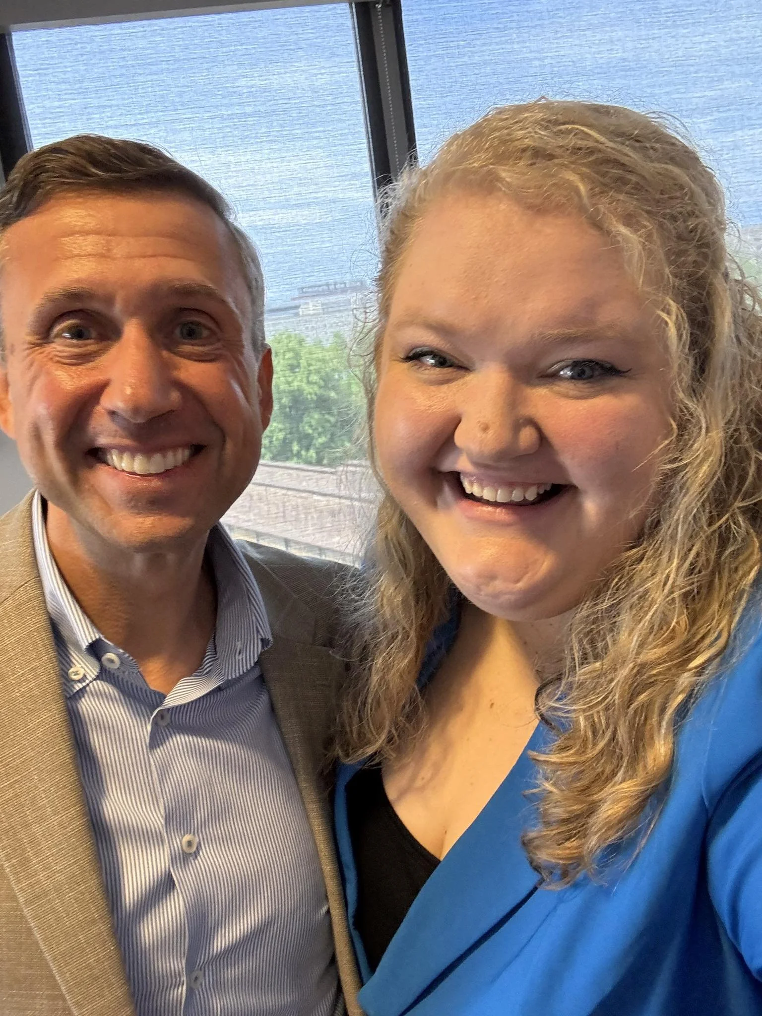 A smiling man and woman taking a selfie indoors near a window with a water body and green trees in the background.