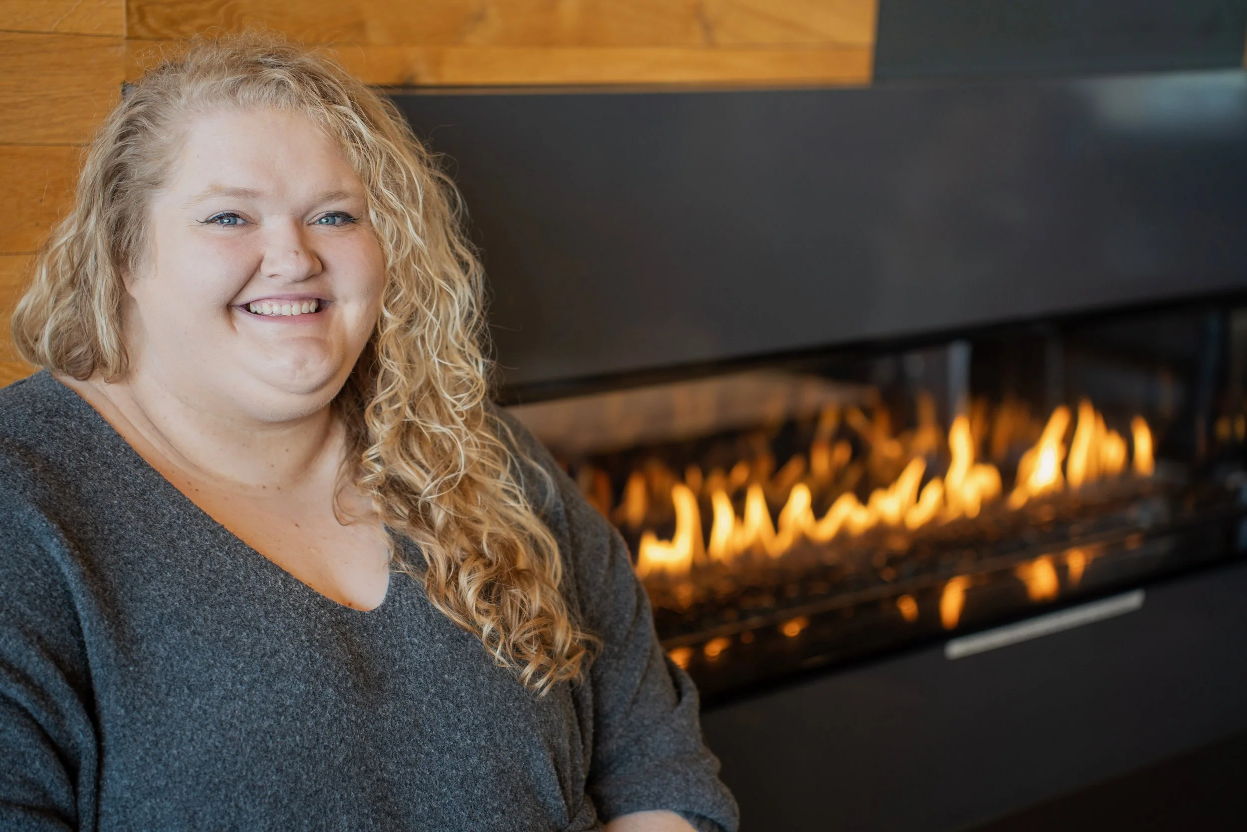 A woman with curly blonde hair smiling while sitting in front of a modern fireplace with visible flames.
