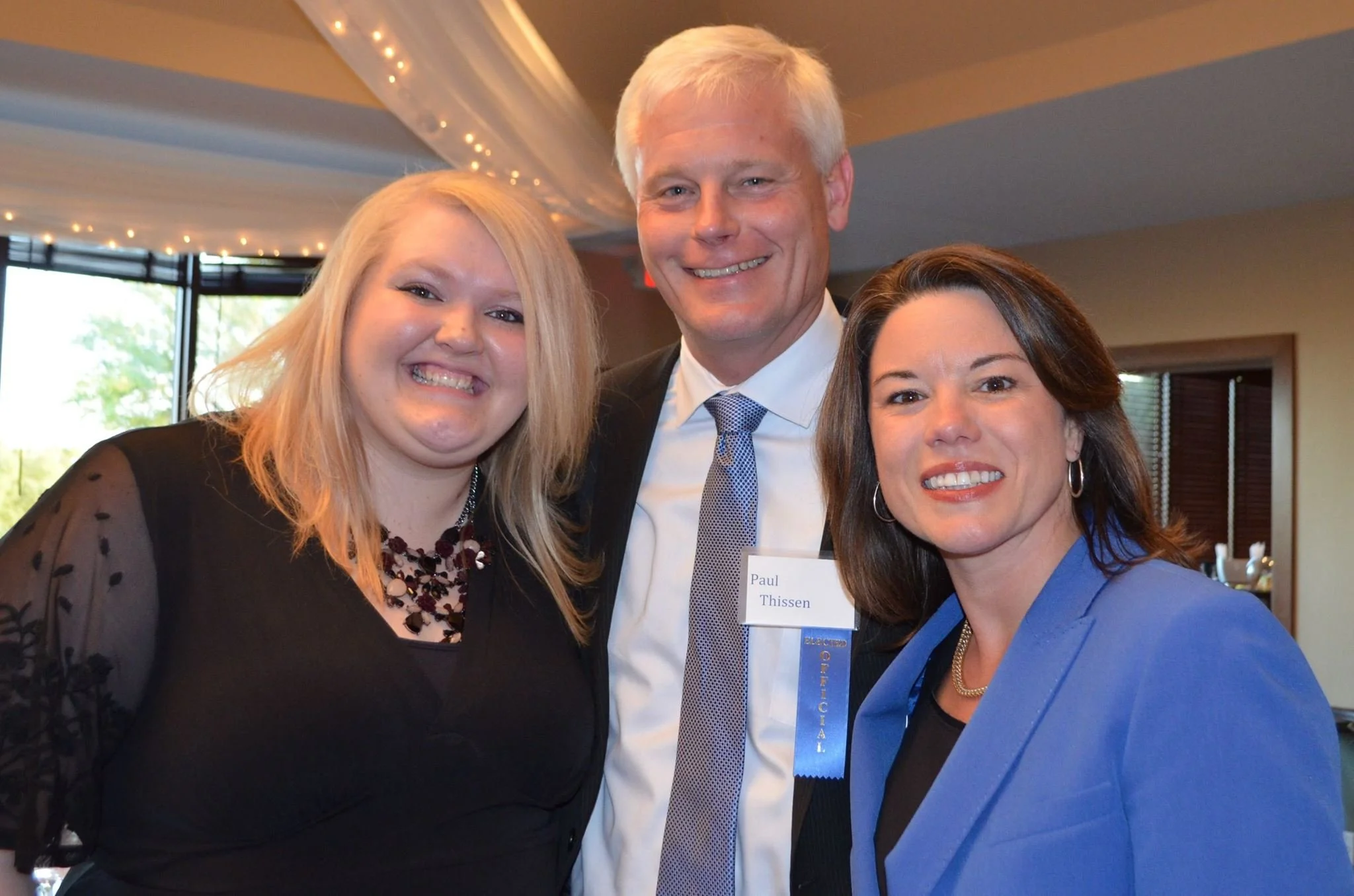 Three smiling people at a professional event, with a woman on the left wearing a black dress, a man in the middle with a name tag that reads 'Paul Thissen' and wearing a suit, and a woman on the right wearing a blue blazer.
