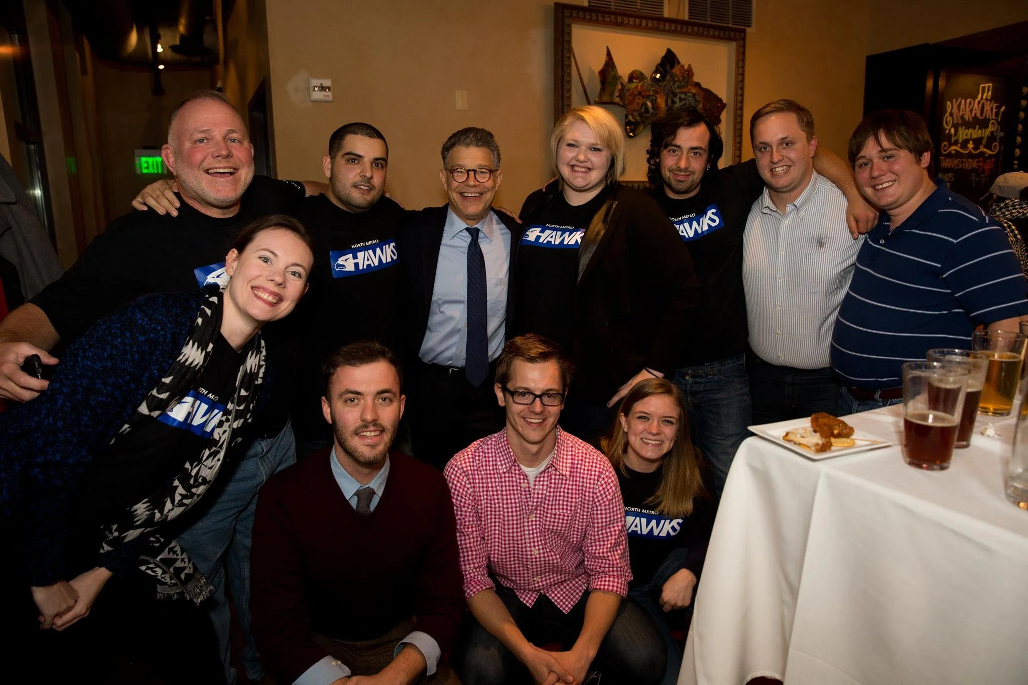 Group of men and women smiling and posing together in a restaurant or banquet hall, with some wearing North Metro Hawks shirts, about to enjoy food and drinks.